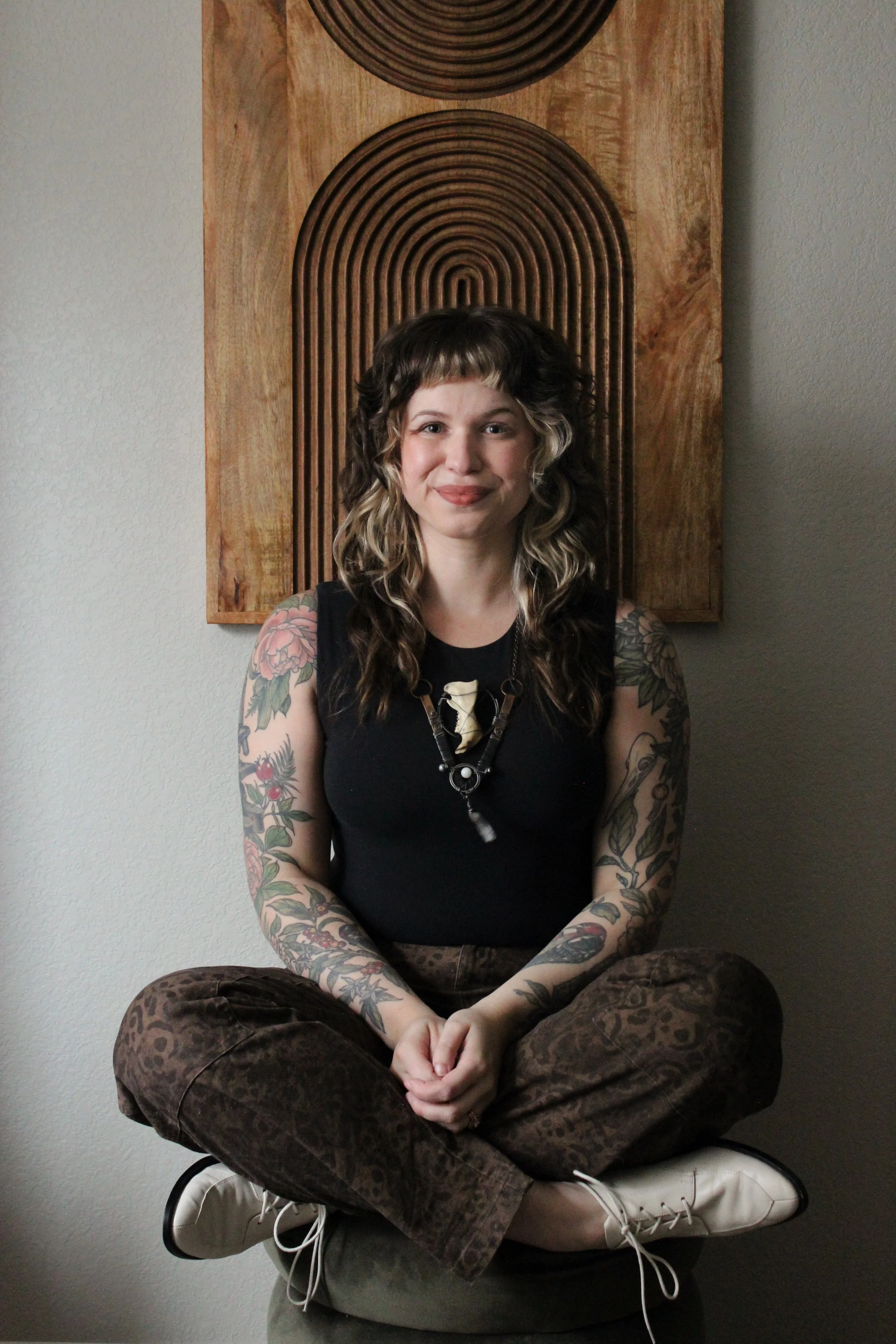 A woman with curly hair and tattoos on her arms and shoulders sitting cross-legged in a relaxed pose in front of a wooden wall art piece with a circular design.