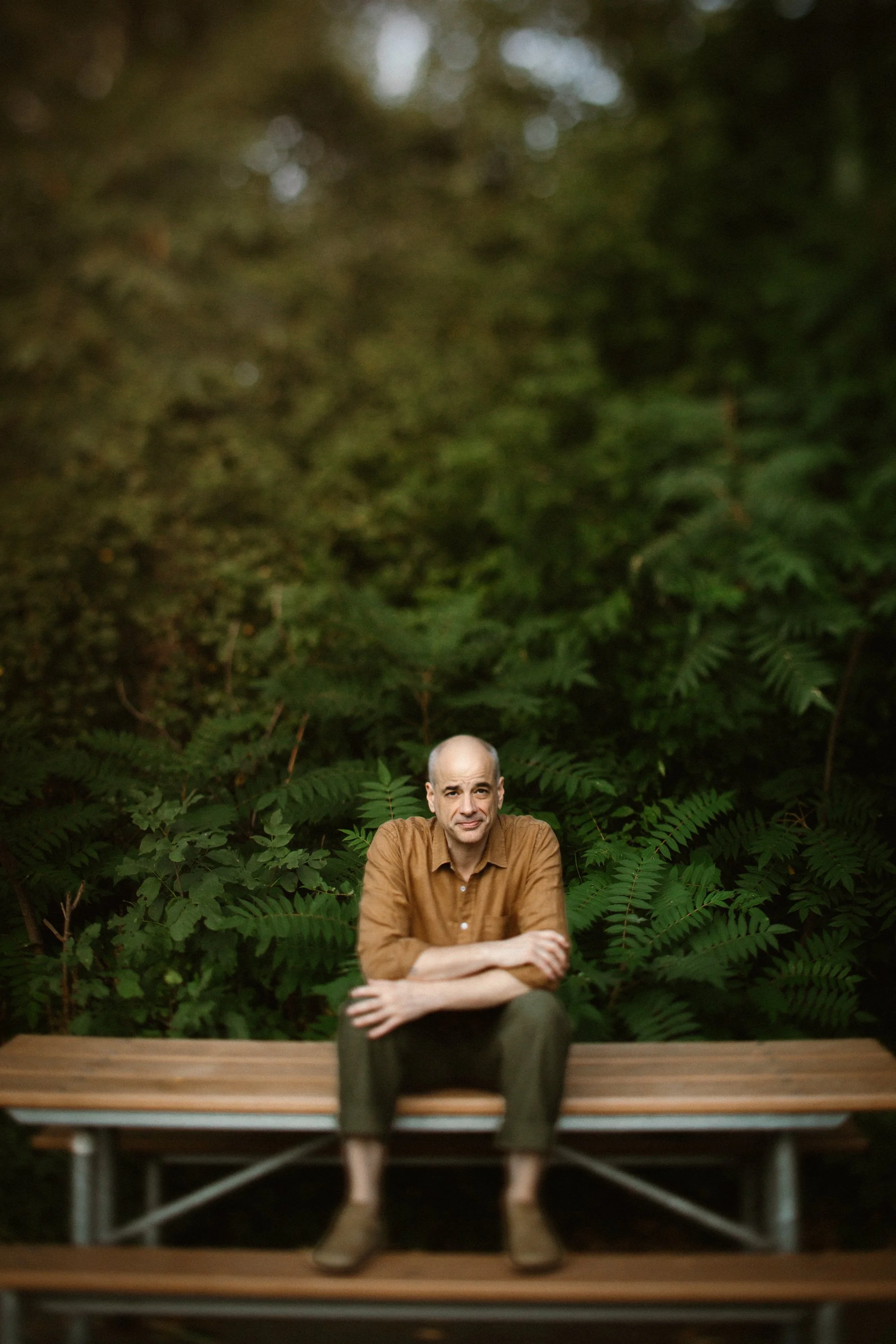 A man sitting on a wooden park bench surrounded by green bushes and plants, with a lush green background.