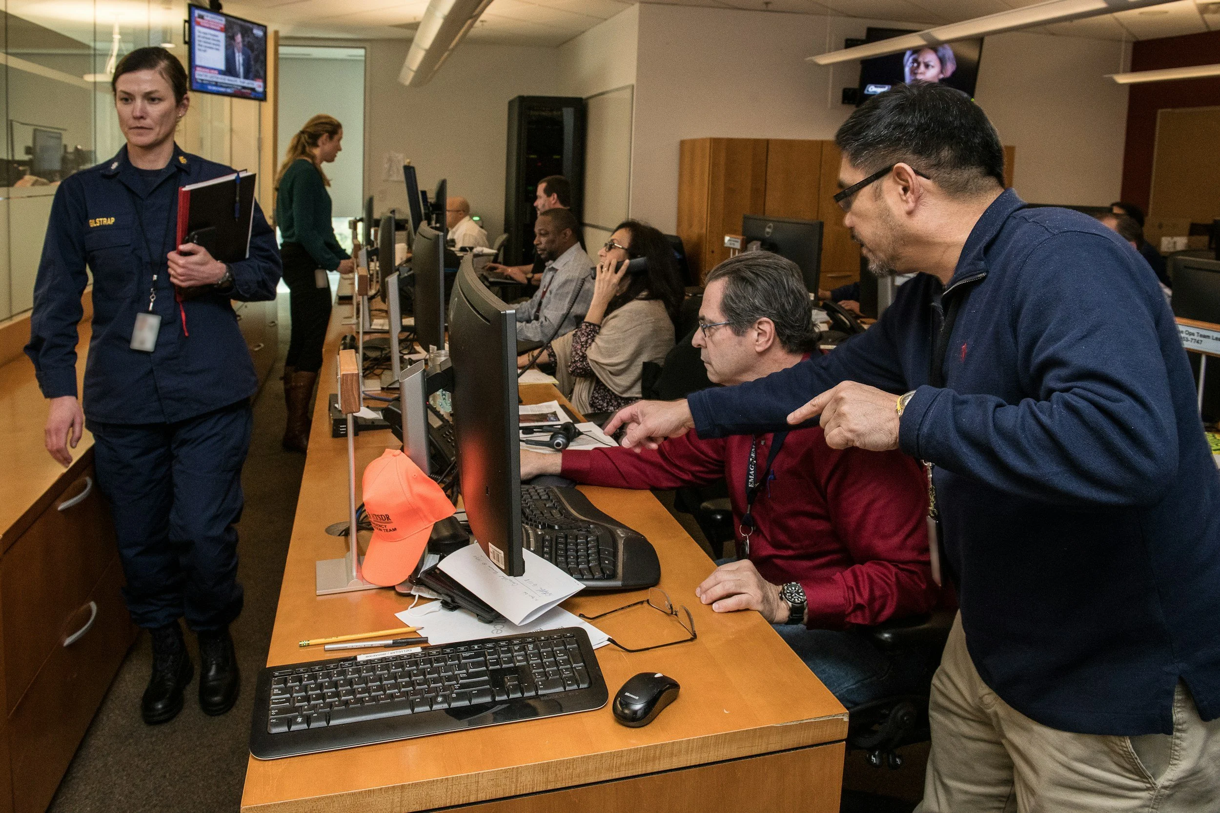 Several people working at computers in a crisis response office, with two individuals standing and one person wearing a navy uniform walking in the foreground.