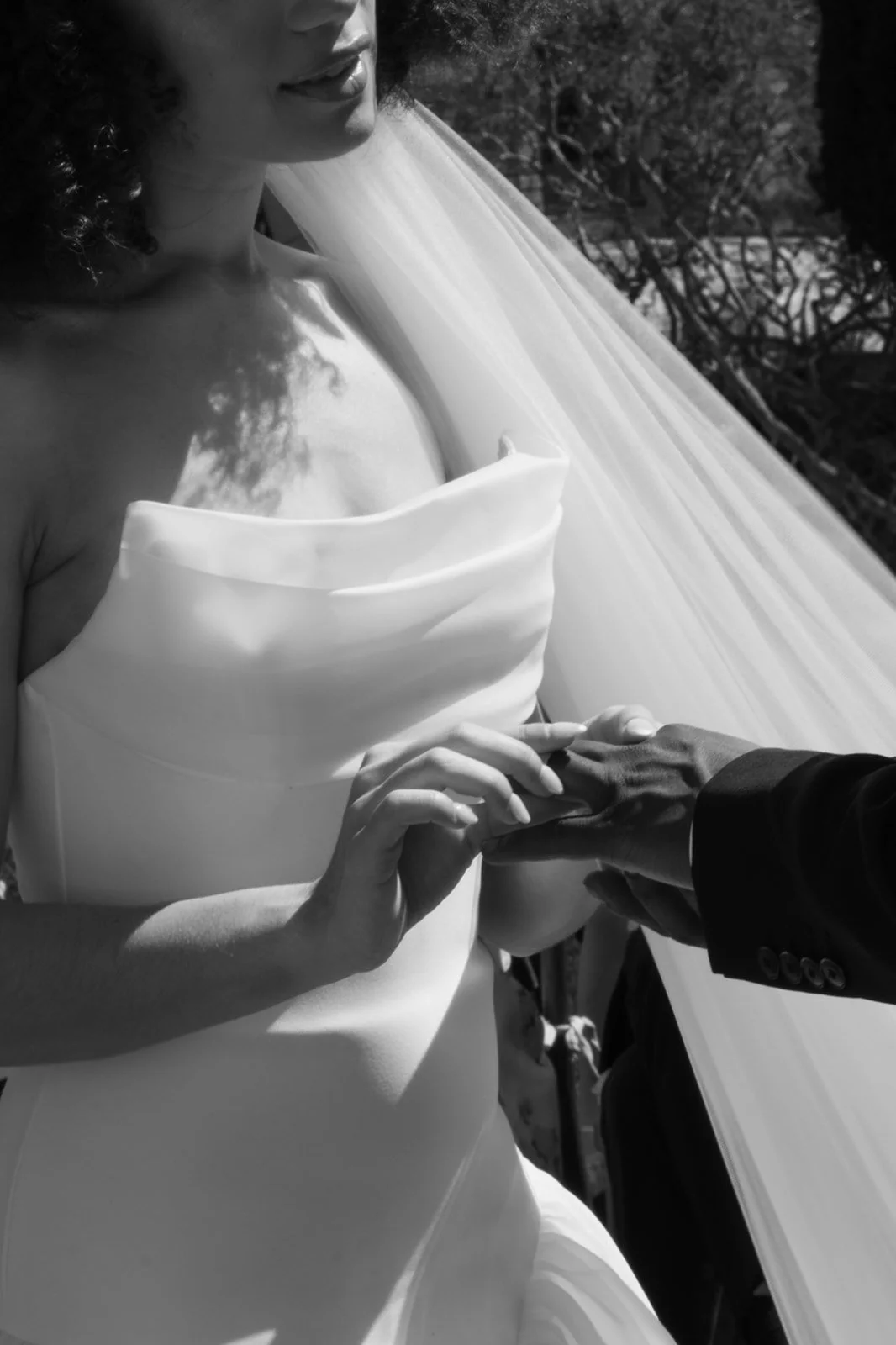 A black and white photo of a bride in a strapless wedding dress, holding hands with someone in a dark suit during a wedding ceremony outdoors.
