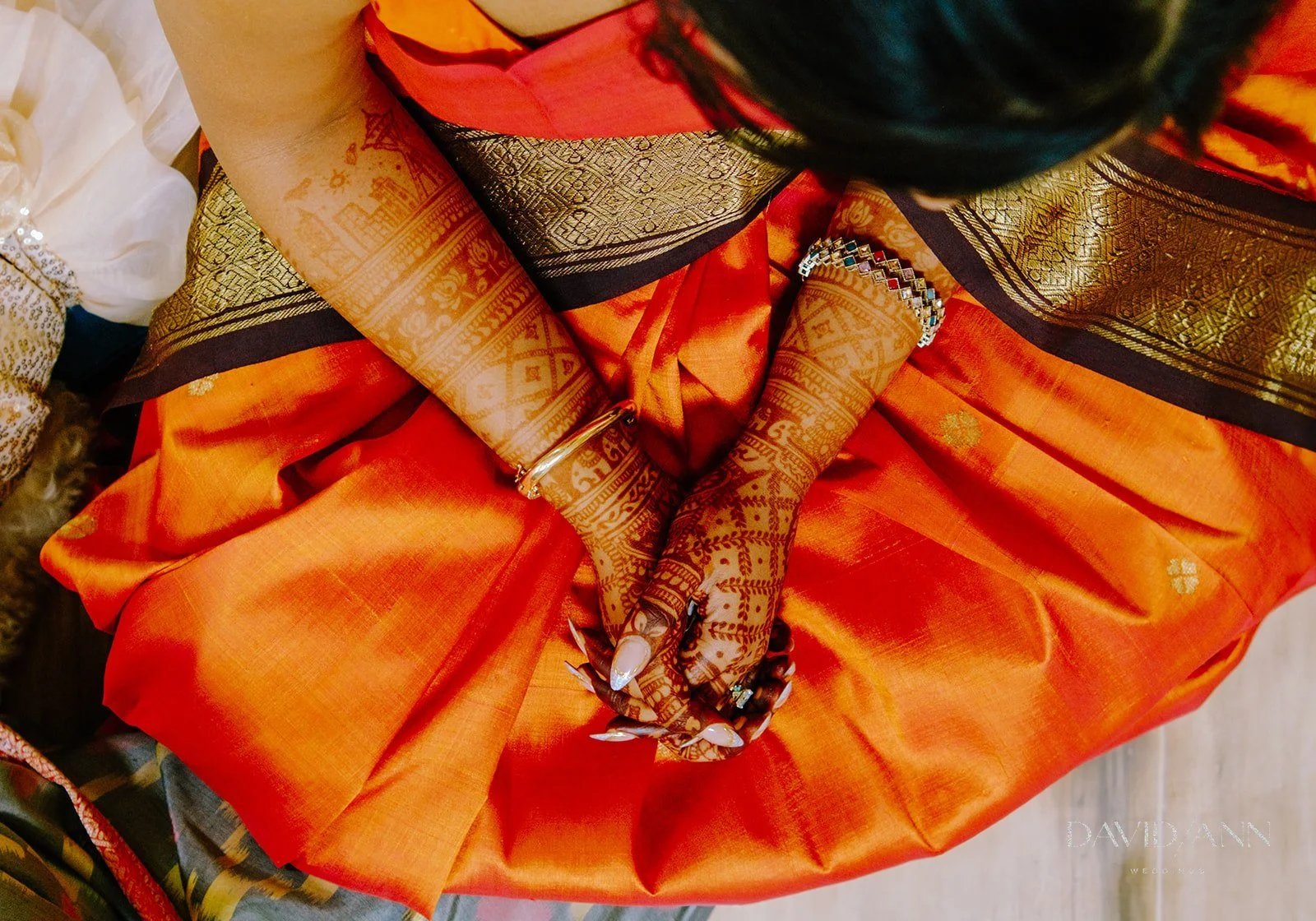 Top-down view of a woman dressed in traditional Indian attire with intricate henna designs on her hands, wearing bangles, rings, and jewelry, sitting with hands folded over a bright orange and black sari.