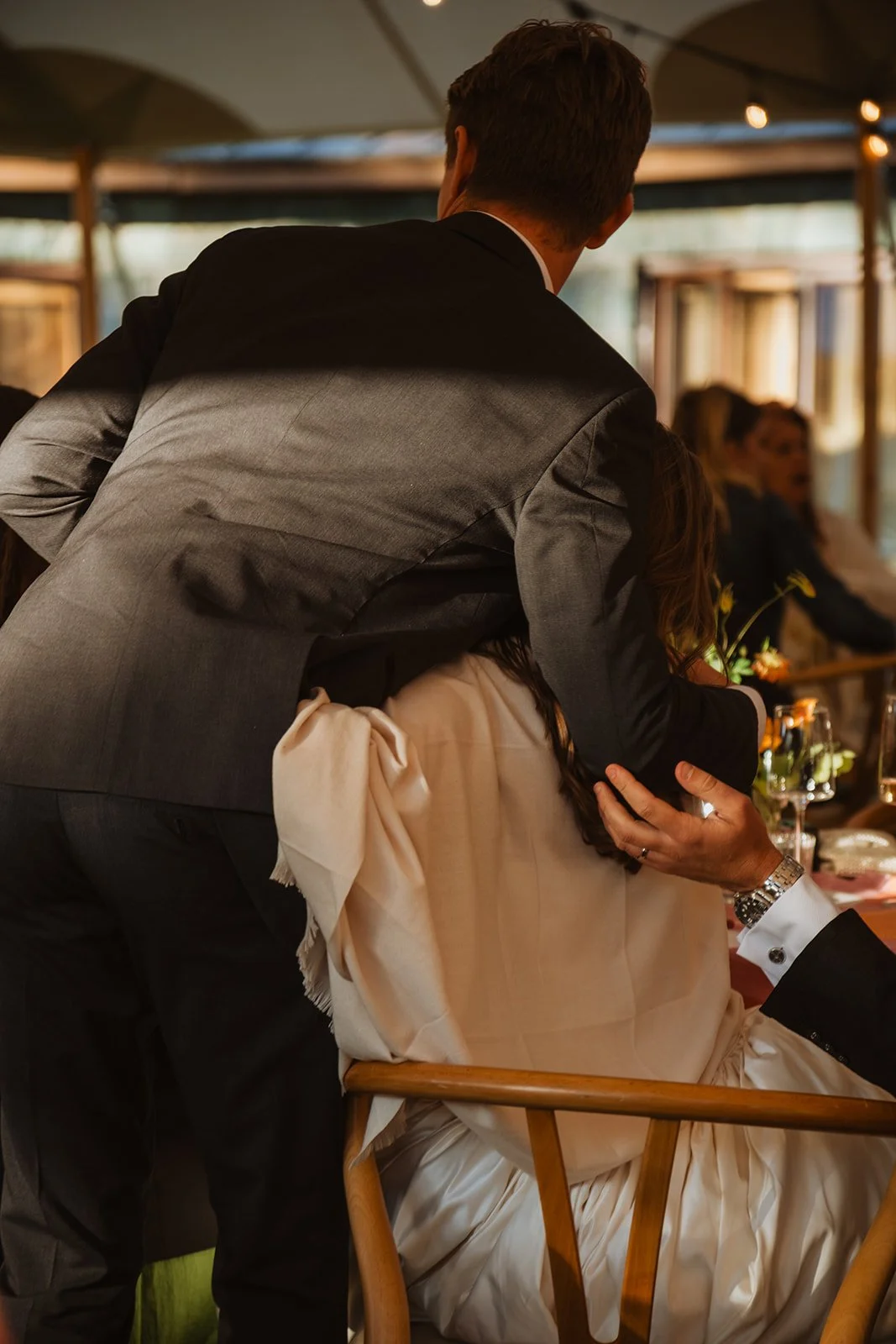 A man in a gray suit hugs the bride sitting on a wooden chair at a wedding reception.
