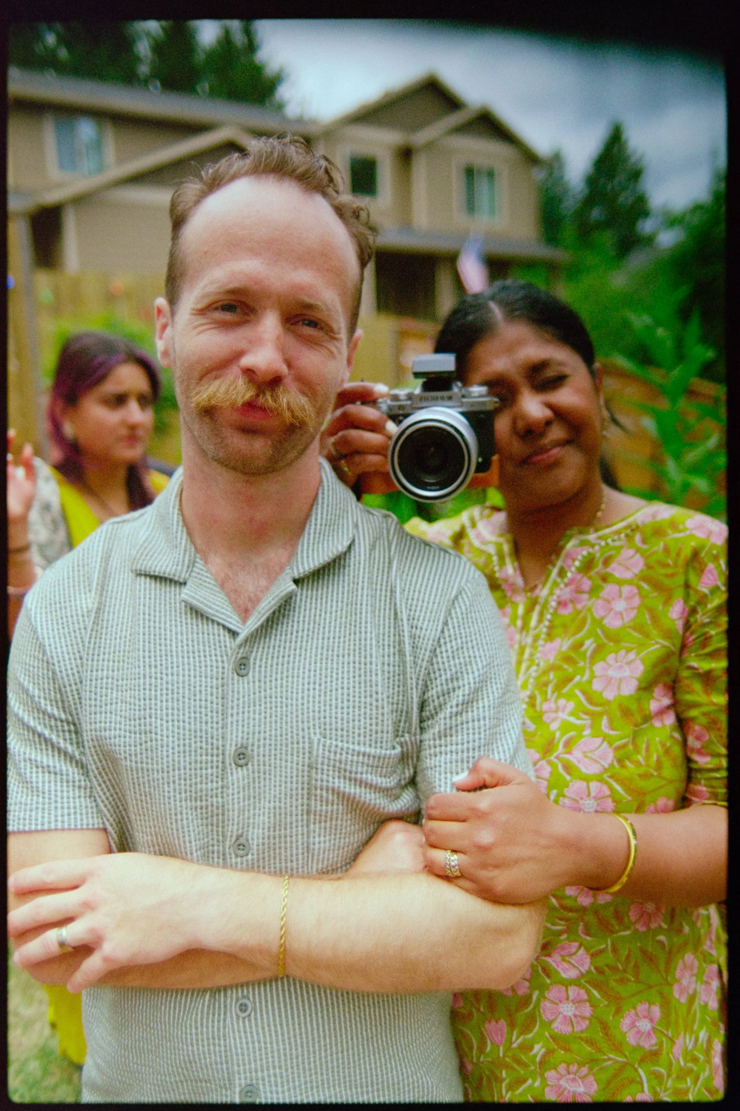 A caucasian man with red hair and mustache smiling and an indian woman behind him with a camera in her hand