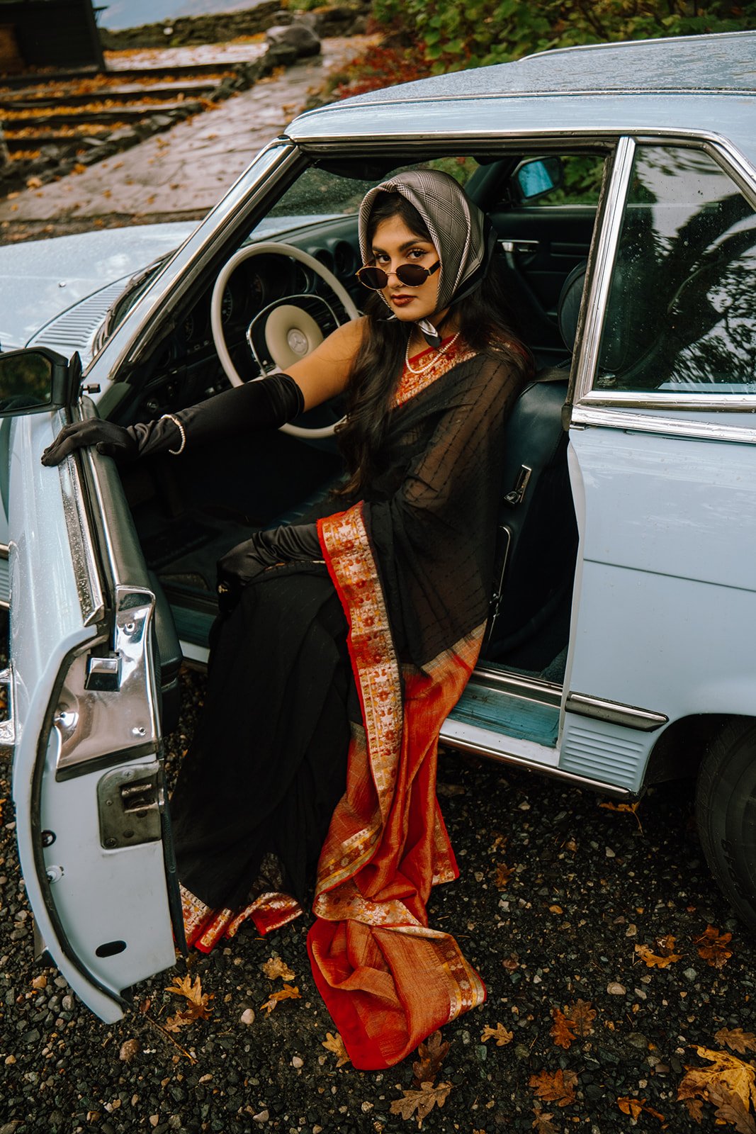 A woman dressed in traditional Indian attire sits in a vintage light blue car with the door open. She is wearing black gloves, sunglasses, a headscarf, and has long dark hair. The background shows autumn leaves and stairs.