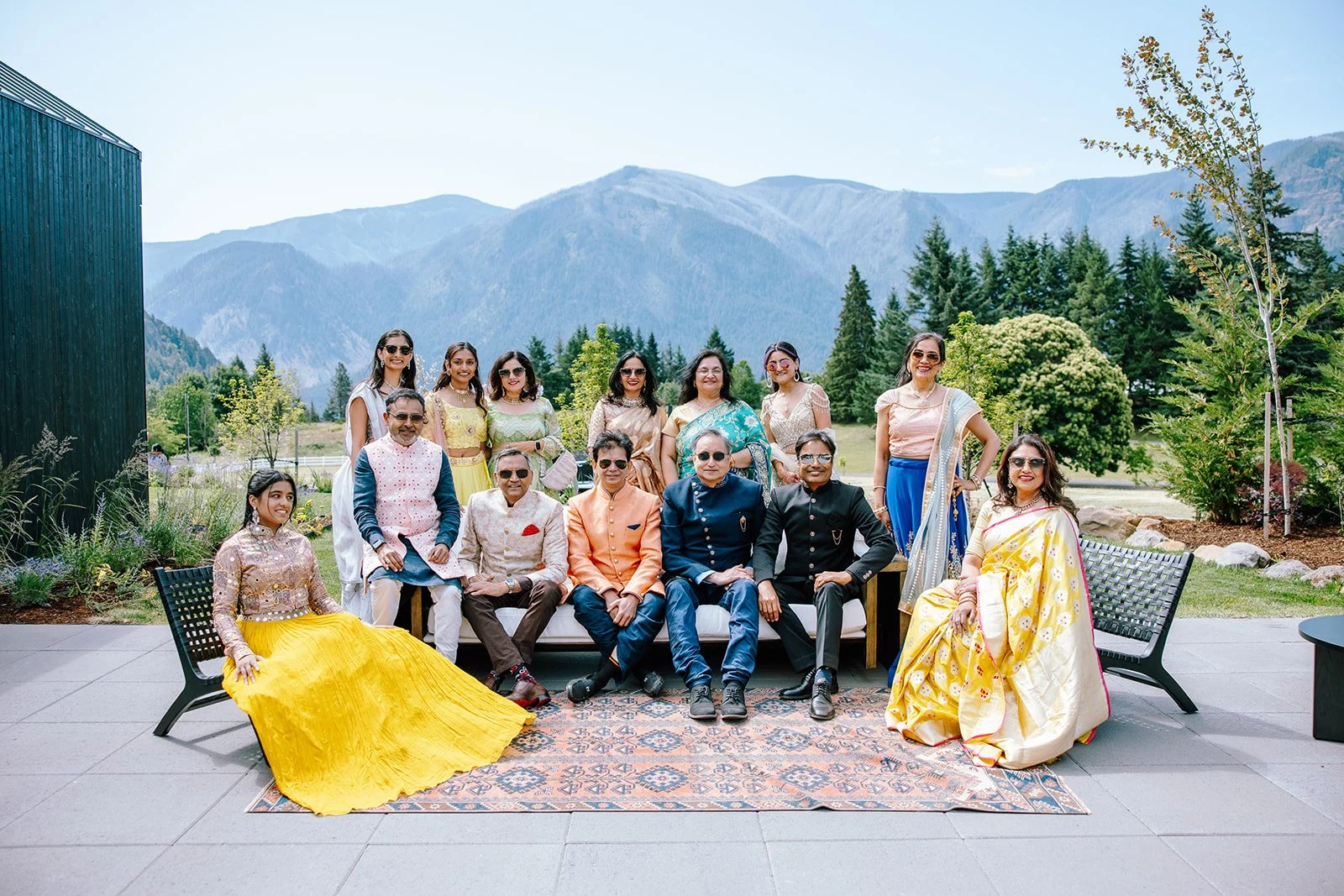 Group of people outdoors, sitting and standing on a patio with mountains, trees, and blue sky in the background. They are dressed in traditional and modern attire, smiling for the photo.
