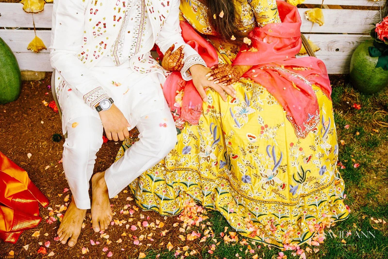 A couple dressed in traditional Indian attire sitting close together outdoors on a flower petal-strewn ground, with a white wooden fence and large pots nearby.