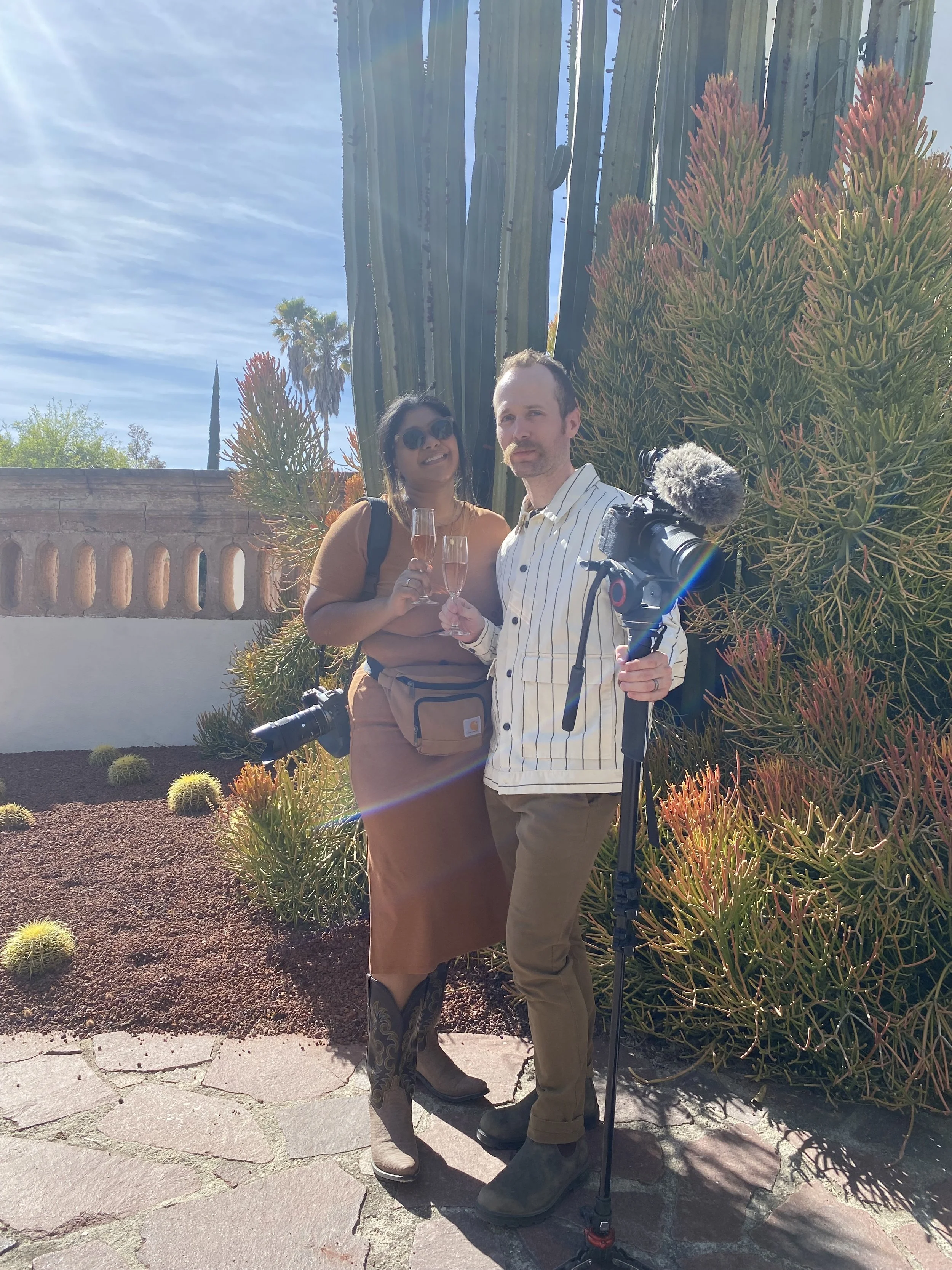 Two people celebrating with champagne glasses in front of a large cactus and succulent plants outdoors on a sunny day.