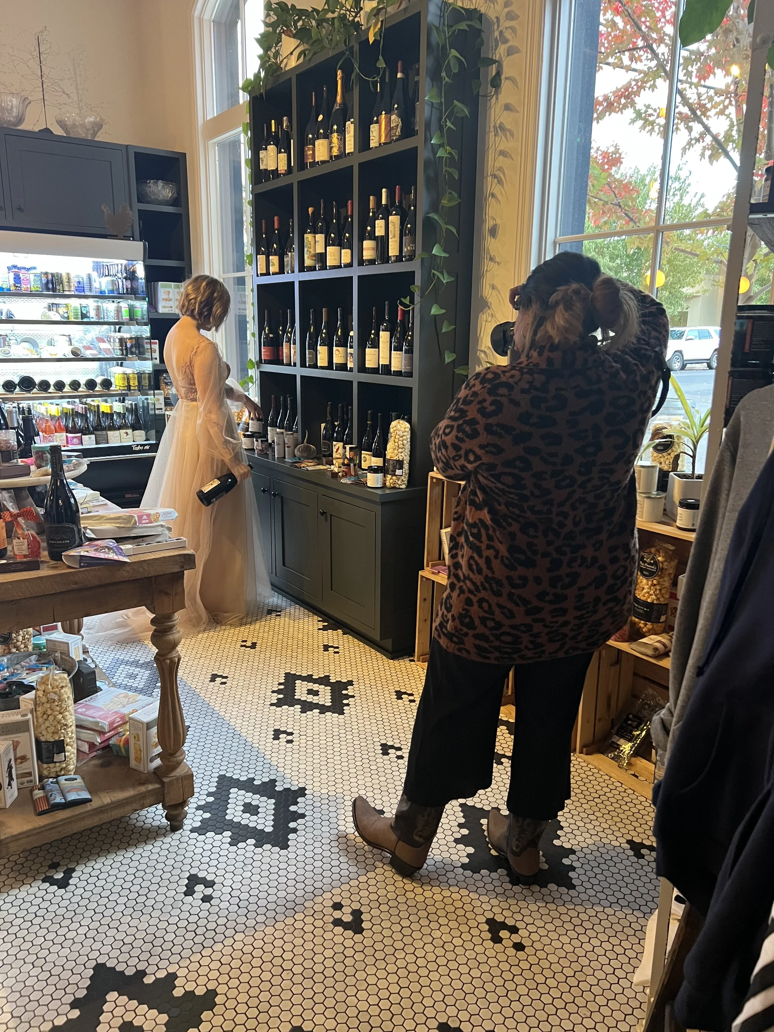 Inside a boutique store with a woman in a light-colored dress looking at wine bottles on a shelf, a photographer taking pictures, and a patterned black and white tile floor.