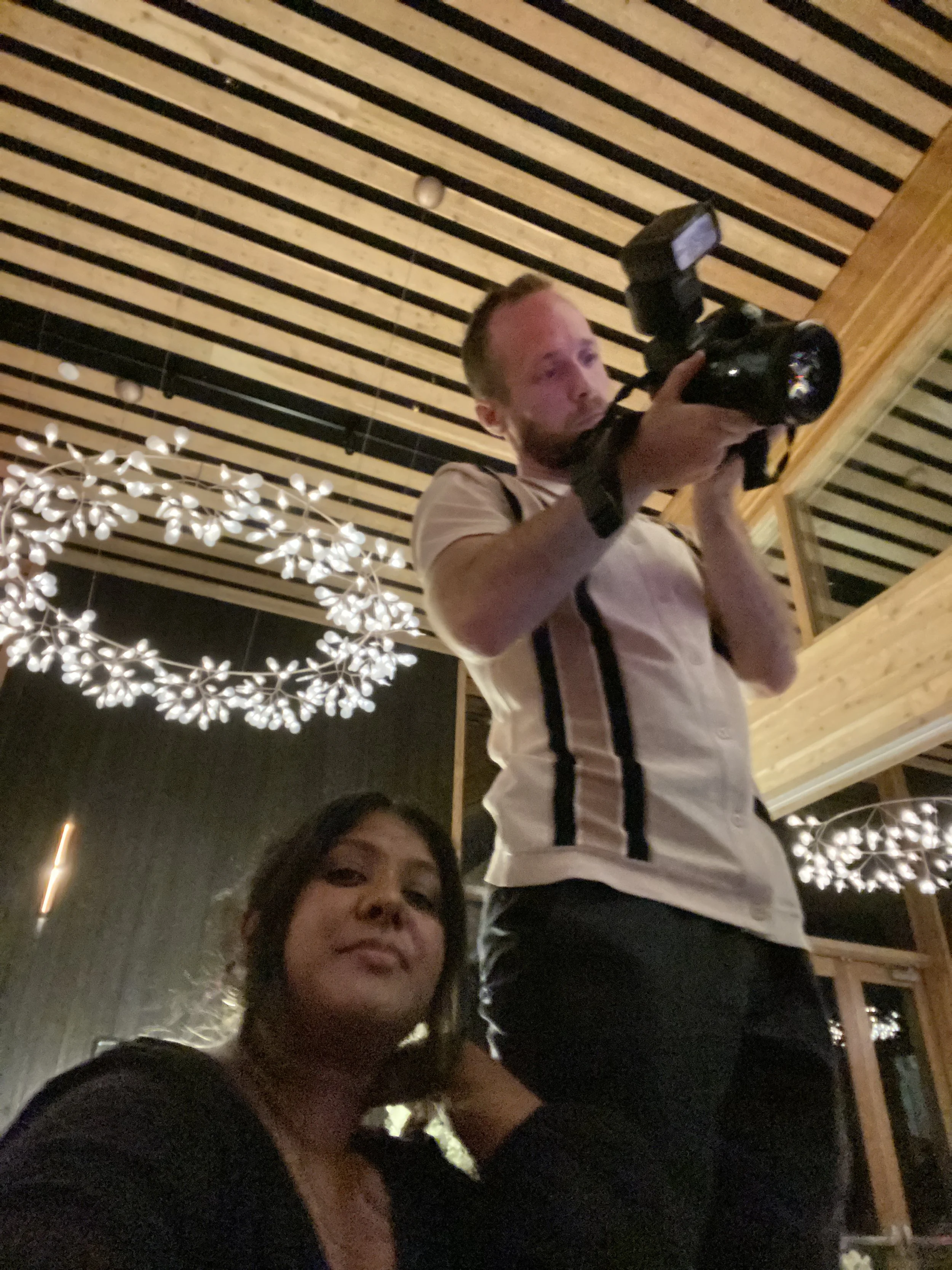 A man holding a camera with an external flash at an indoor event, looking through the viewfinder, and a woman sitting in front of him, smiling at the camera.