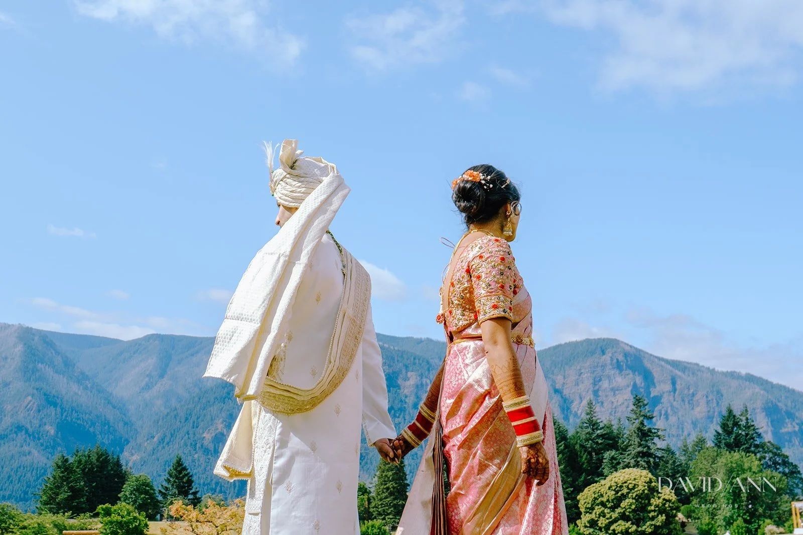 A bride and groom holding hands outdoors with mountains and trees in the background.