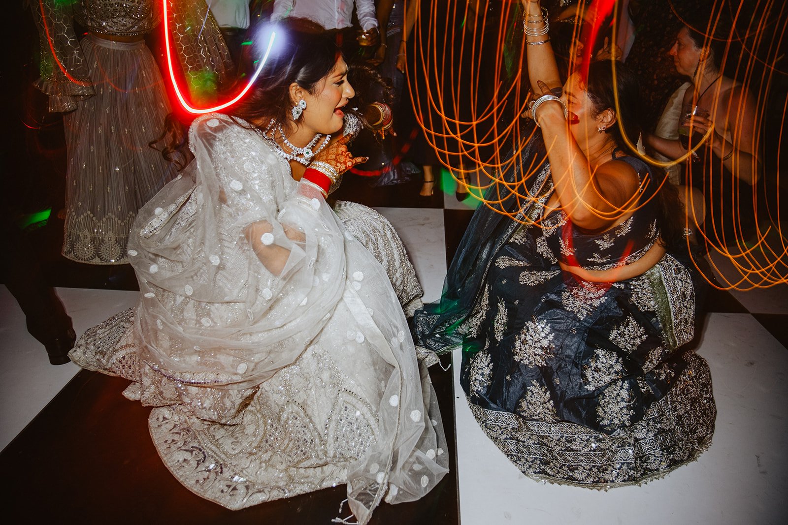 Two women dressed in traditional South Asian attire dance and celebrate at a festive event, with colorful lights and a checkered floor in the background.