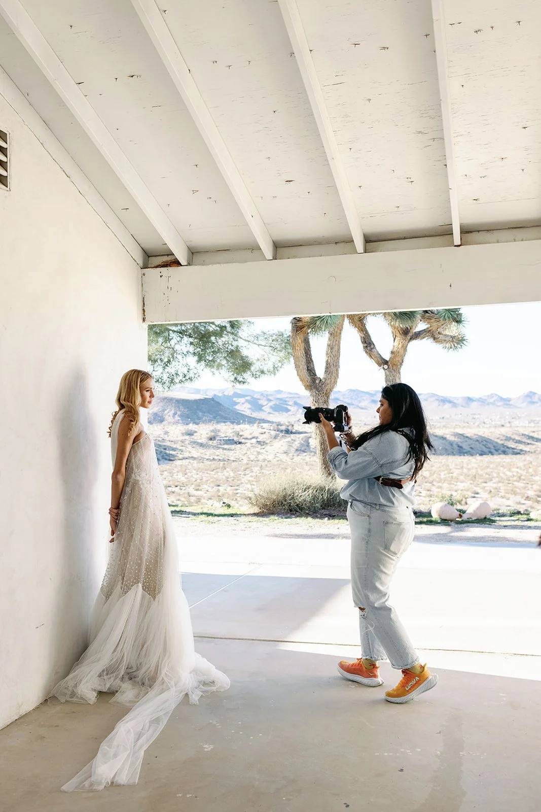 A woman is taking a photo of a woman in a wedding dress under a covered porch with desert landscape and mountains in the background.