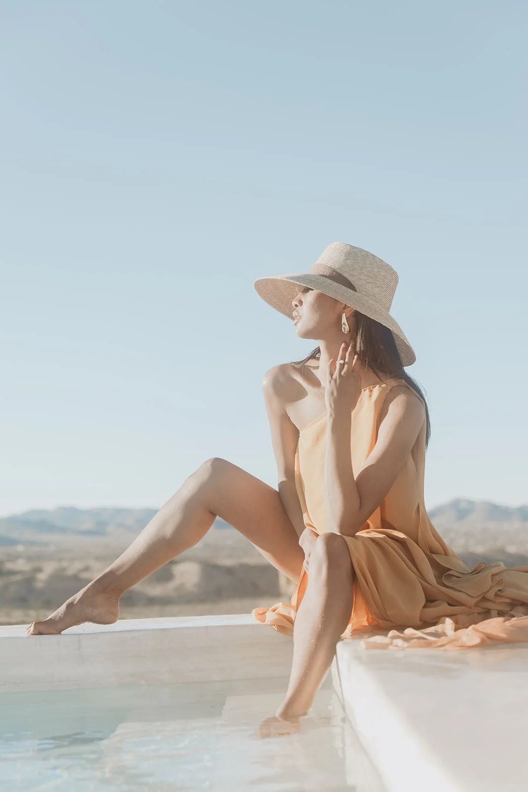 Woman sitting by a pool wearing a large sun hat, earrings, and a peach-colored dress, with a desert landscape in the background.