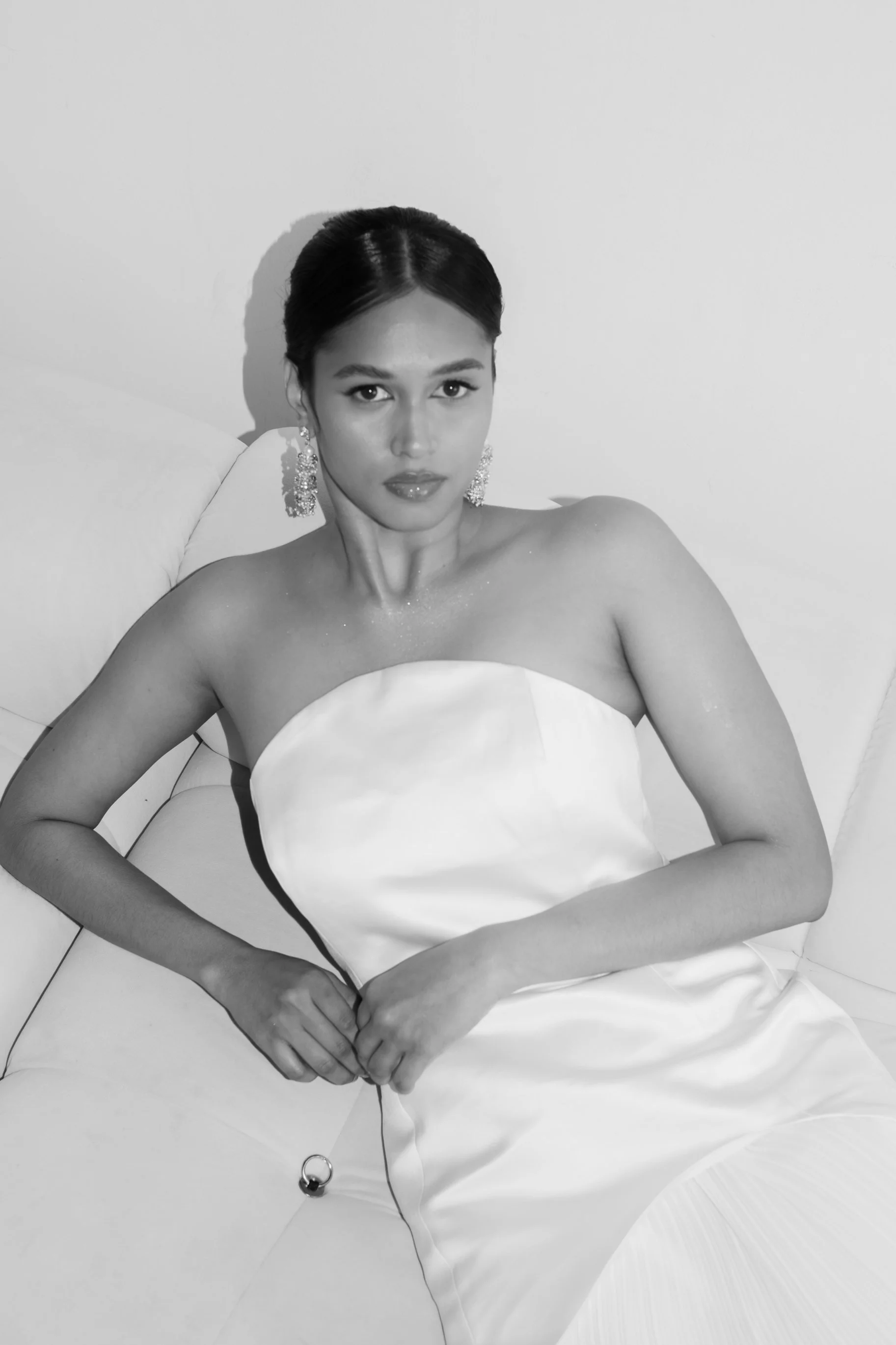 Black and white editorial photo of an indian woman in a strapless dress with earrings, sitting on a sofa, looking directly at the camera.
