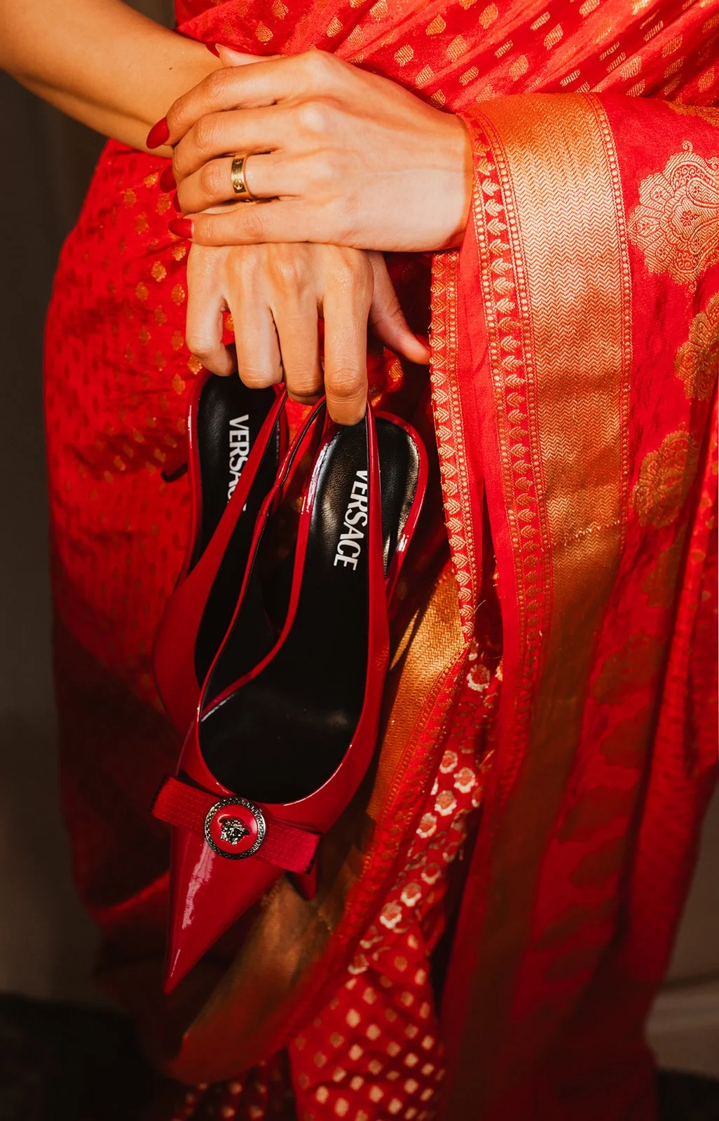 Person holding red Versace high-heeled shoes, dressed in a red and gold embroidered saree.