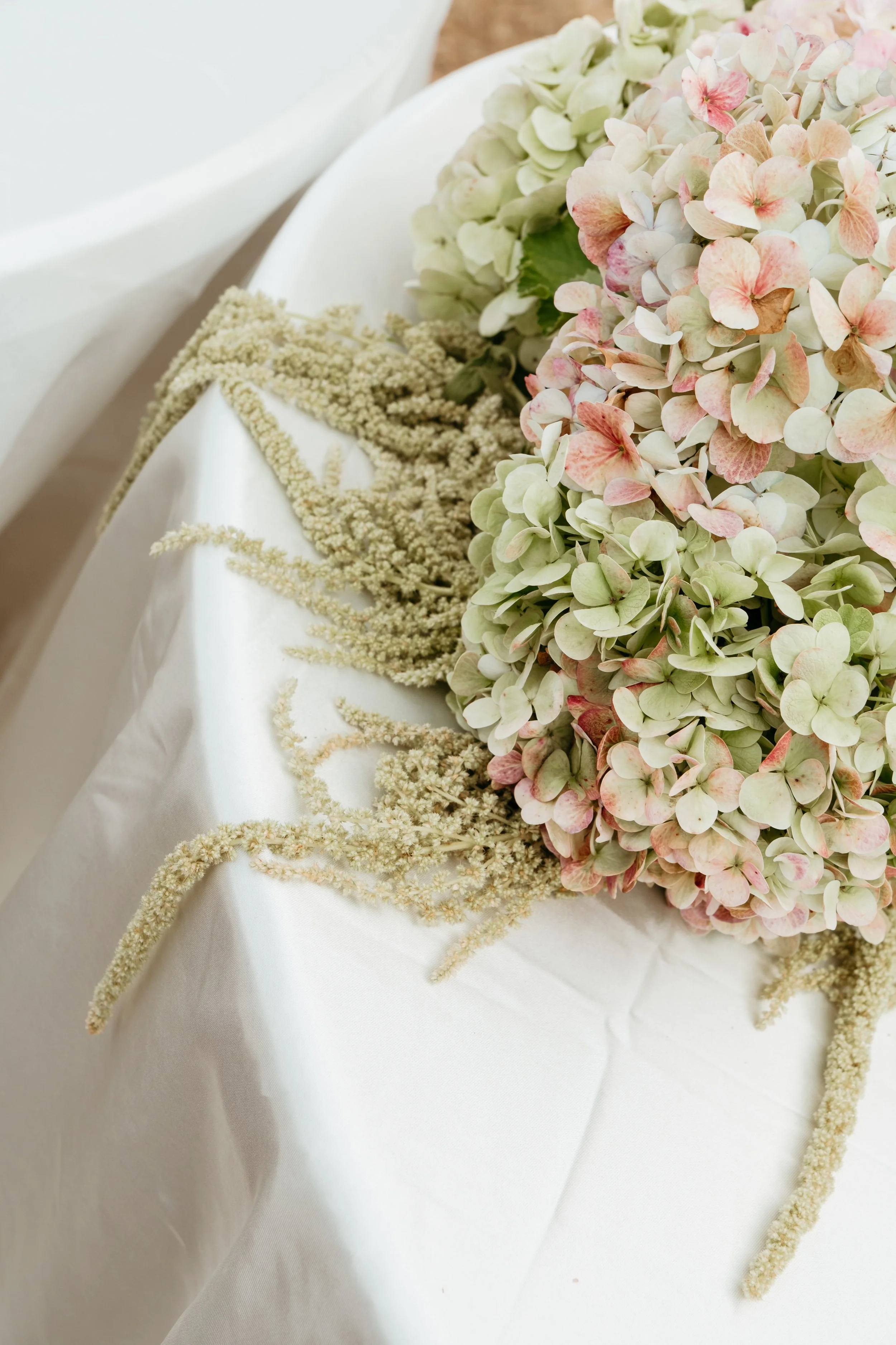 A bouquet of pastel pink and green hydrangea flowers on a white satin-covered table with white floral accents.