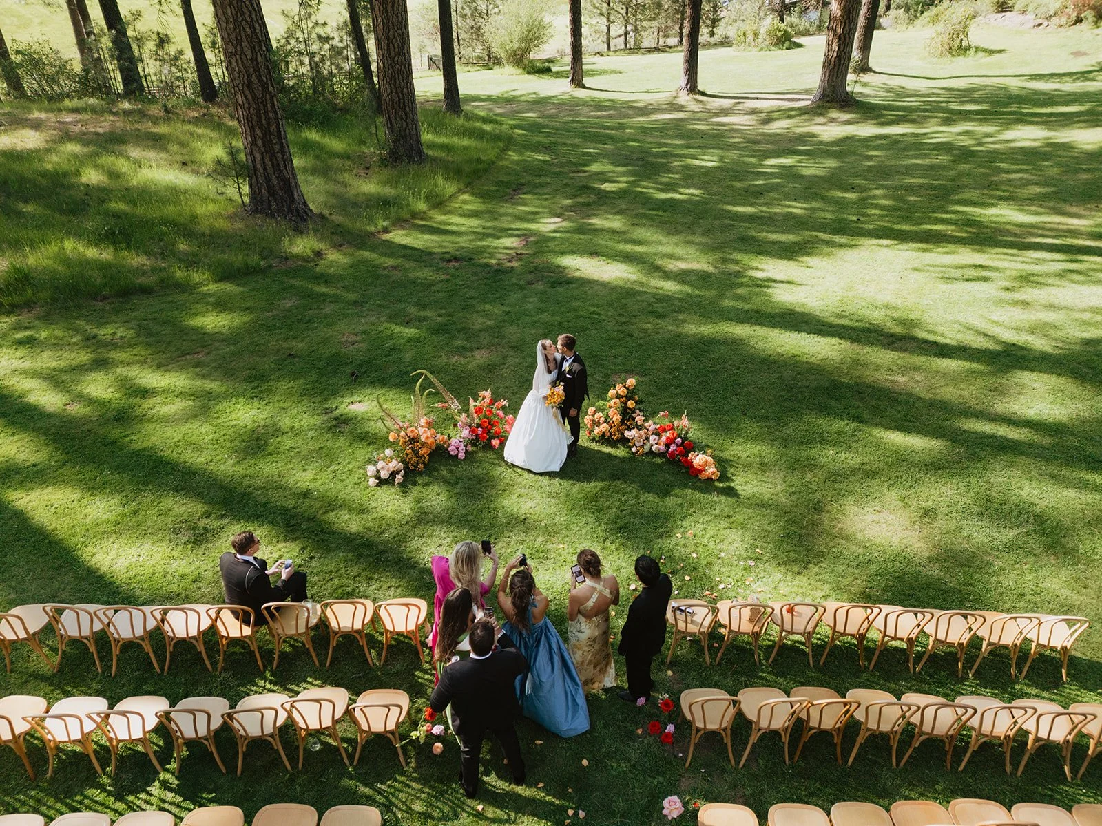 A wedding ceremony taking place outdoors in a green, wooded area. The bride and groom are kissing in front of a floral arrangement, with guests seated and standing nearby, some taking photos.