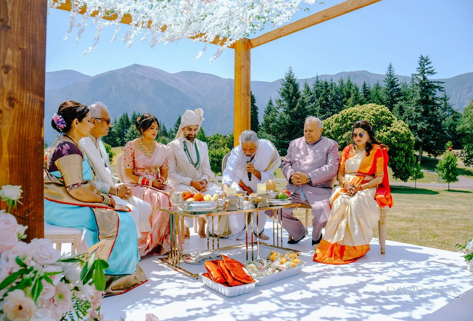 Hindu wedding ceremony outdoors with a group of people, including men and women in traditional attire, seated under a decorated canopy with mountains and trees in the background.