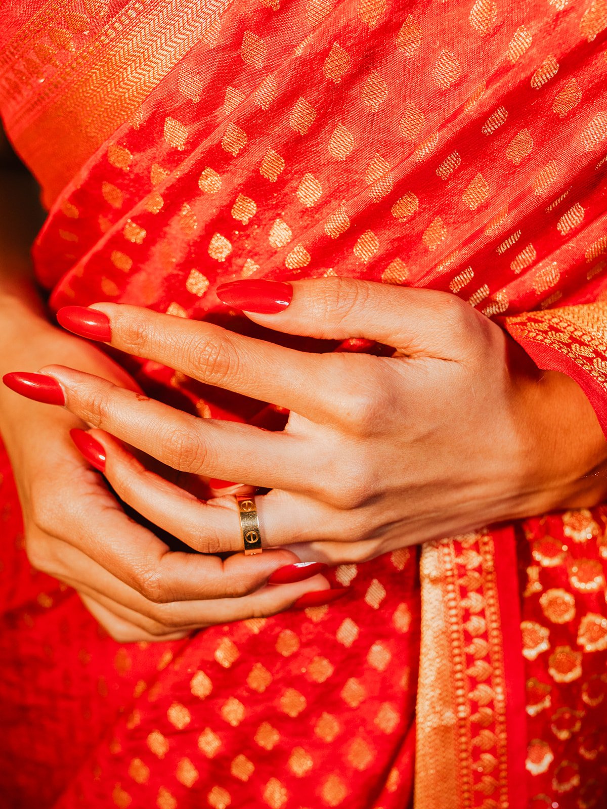 Close-up of a woman's hands with red painted nails and a gold ring, resting on her lap covered in a red and gold traditional Indian fabric with intricate patterns.