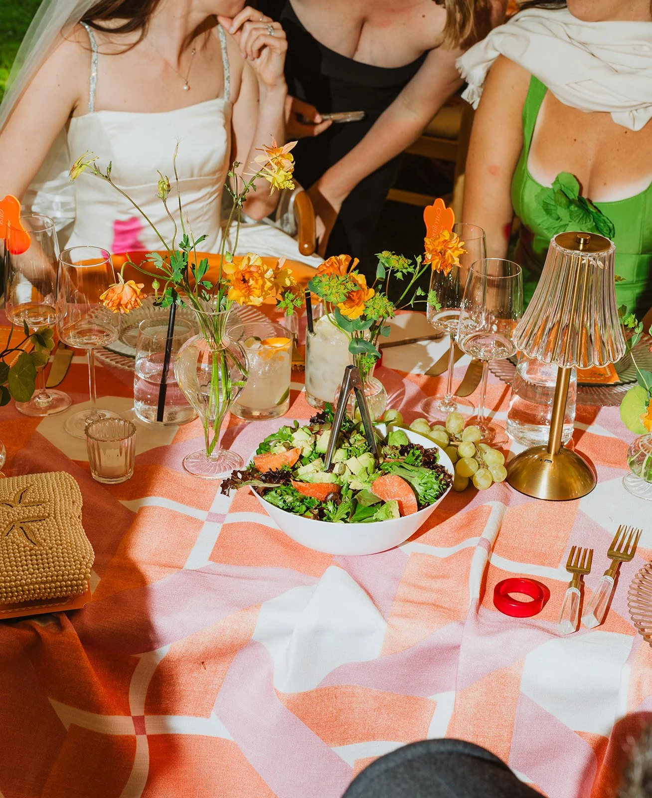 A decorated table at a social gathering with glasses, flowers, salad, and a salad bowl. People are sitting and standing around the table, partially visible.