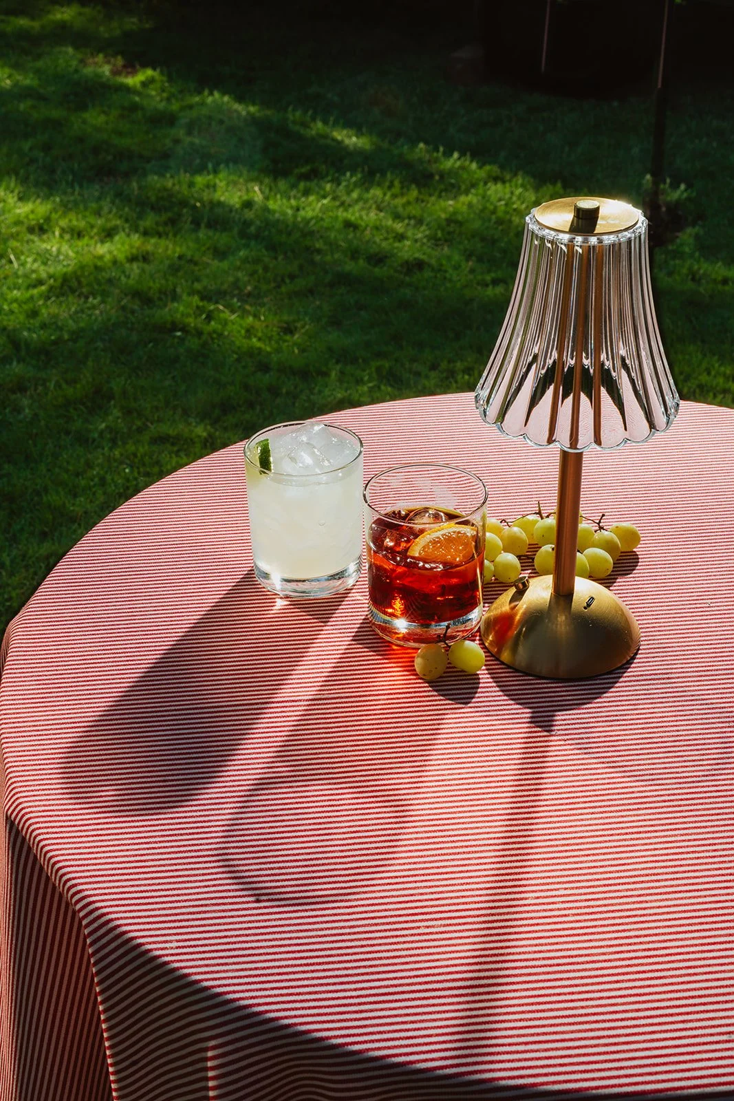 A round table with a red and white striped tablecloth holding two glasses of drinks, a gold table lamp, and bunches of green grapes outdoors on grass.