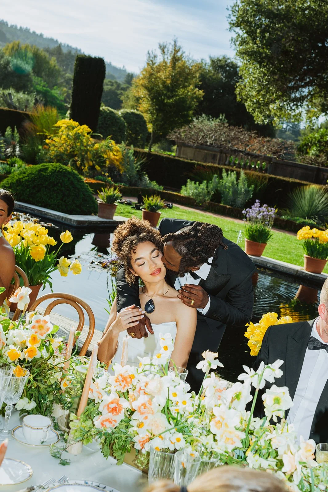 Groom in a tuxedo kissing his wife in a white strapless gown from Besa bridal, with an outdoor garden and pond with yellow flowers in the background, at their wedding banquet in Filoli Gardens in California.