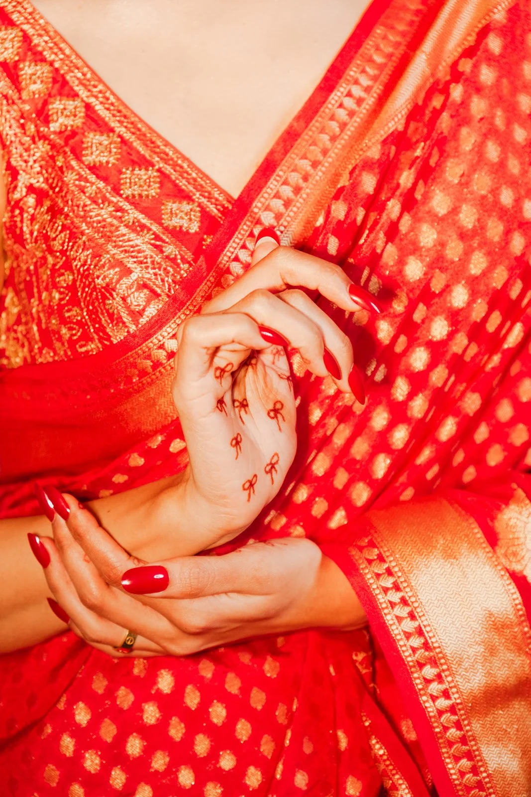 Close-up of a woman in a red traditional saree, with matching red nail polish, holding her hand near her chest, showing small red bow tattoos on her hand and wrist.