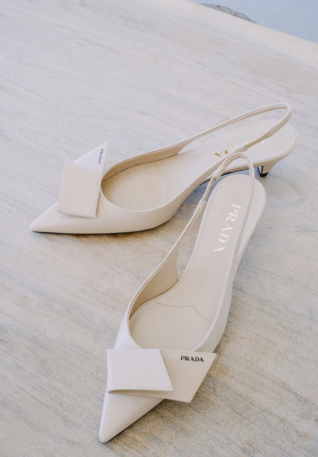 Pair of white Prada high-heeled shoes with pointed toes and decorative bows on a light-colored surface.