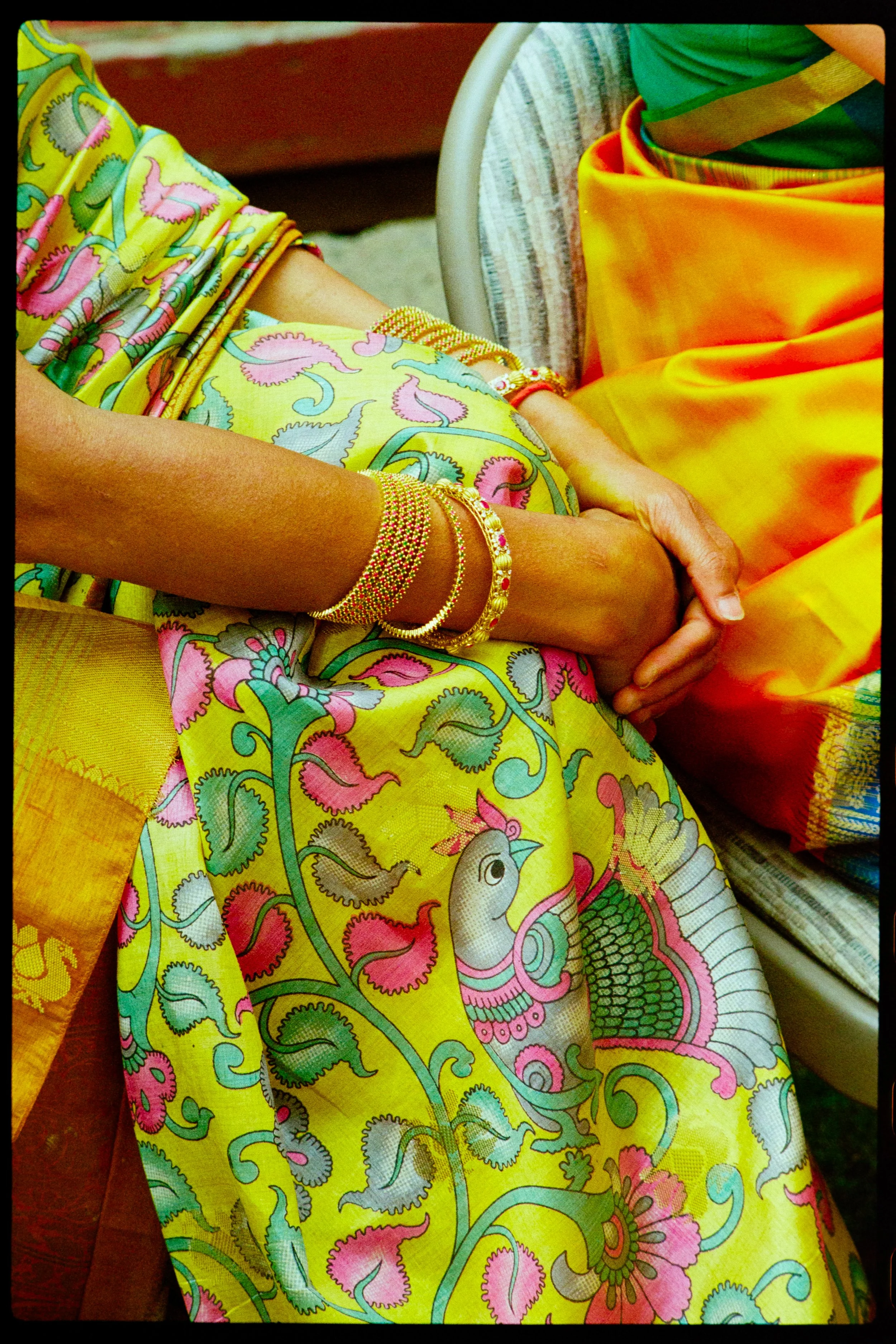 Film photo of two women dressed in vibrant traditional Indian sarees with intricate patterns, sitting together with hands clasped, wearing gold jewelry.