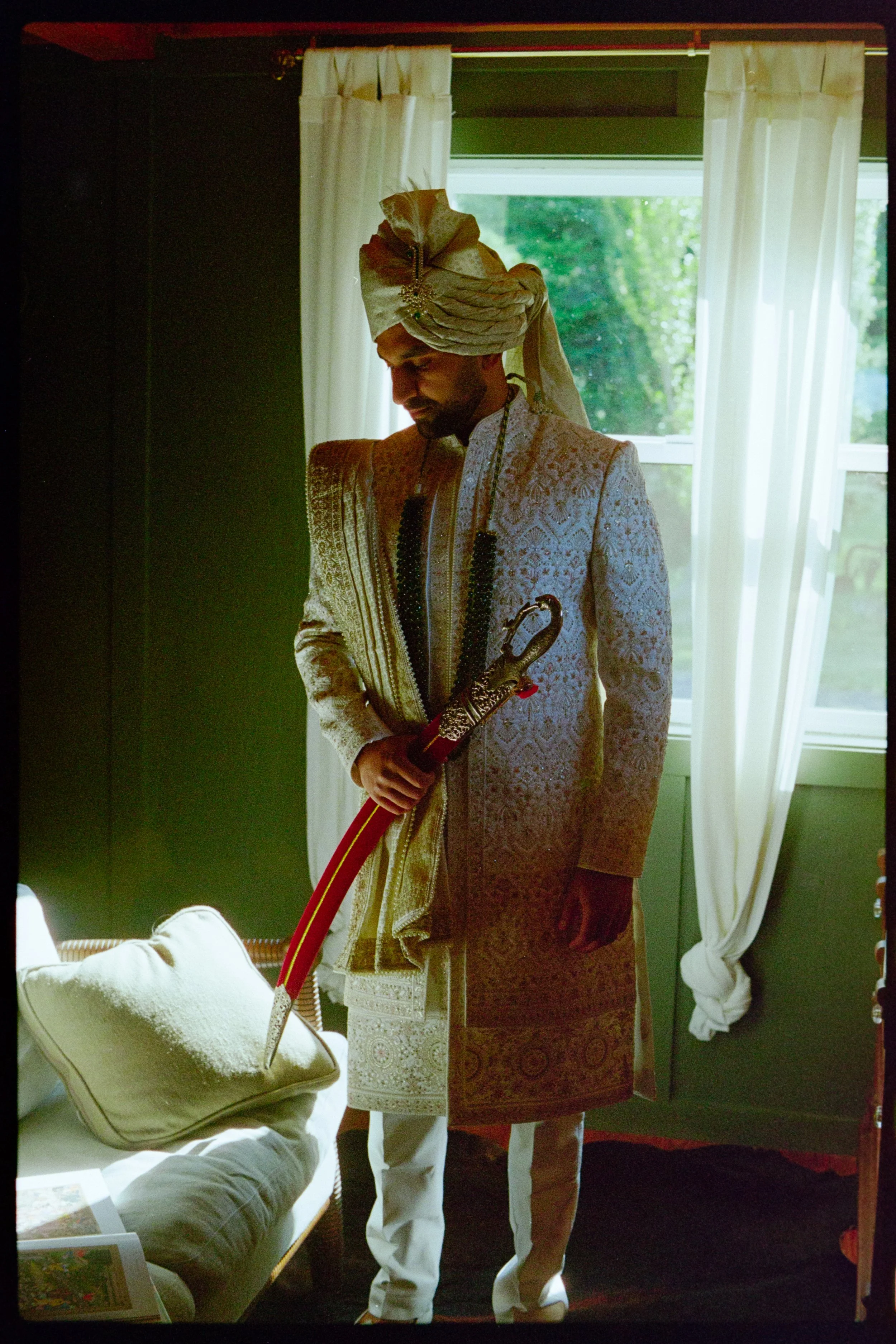 An Indian groom Idressed in traditional Indian wedding sherwani suit, wearing an ornate cream-colored sherwani and turban, holding a decorative sword inside a room with a window and white curtains.