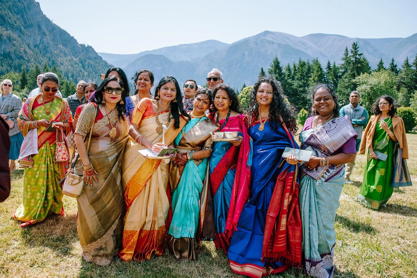 Group of women in colorful traditional sarees standing outdoors in front of mountains and trees.