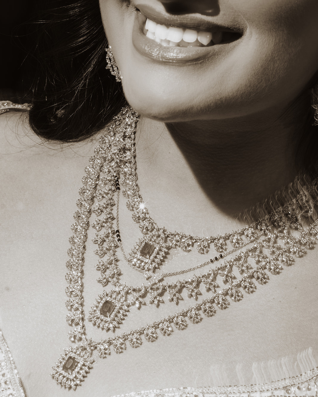 Close-up of an Indian bride smiling, showing her teeth, wearing multiple diamond necklaces and earrings in a black and white photograph.