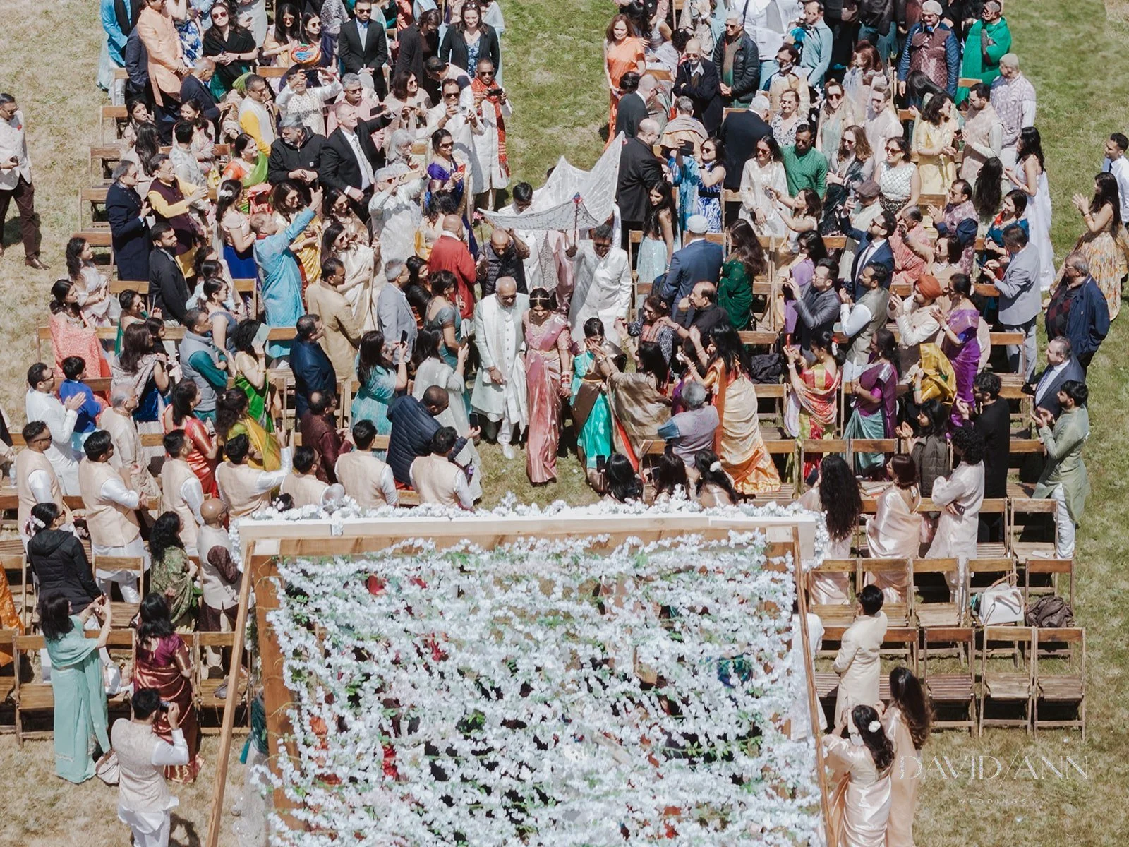 A large gathering of people dressed in traditional Indian clothing at an outdoor wedding ceremony. The scene includes rows of chairs, a decorated canopy, and guests in colorful sarees and suits.