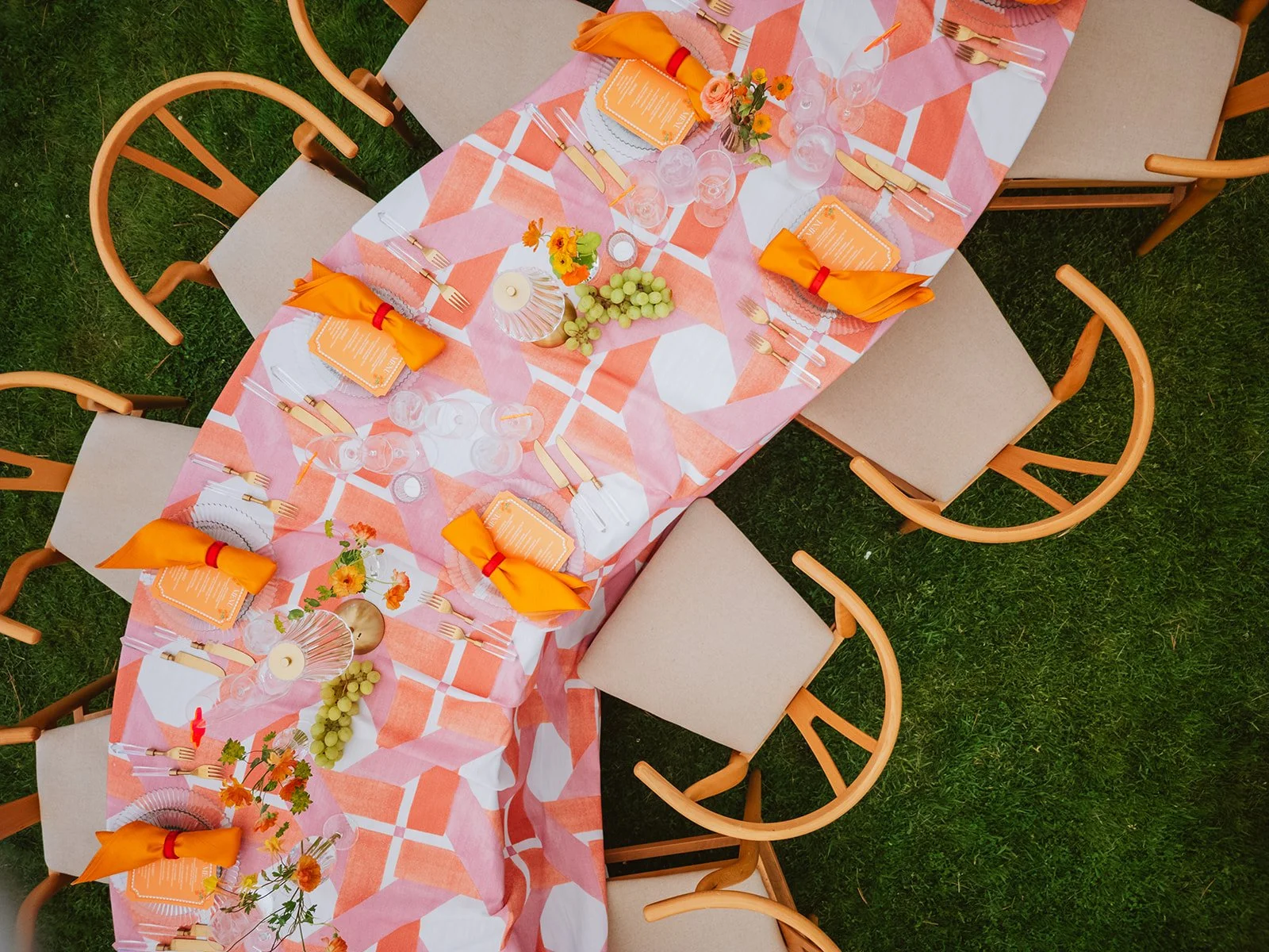 Serpentine styled dining table set for a wedding with pink, orange, and white patterned tablecloth, orange napkins with red bands, clear glassware, cutlery, floral centerpieces, and decorative accents on a grassy area.