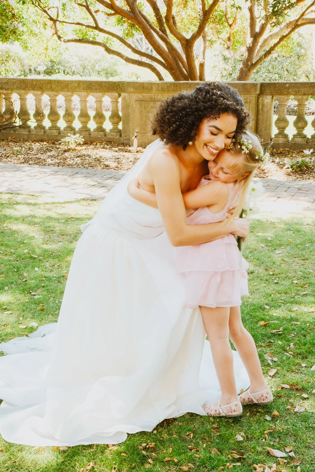 A bride in a white dress hugging her flower girl in a pink dress outdoors on a grassy area, with trees and a stone railing in the background.