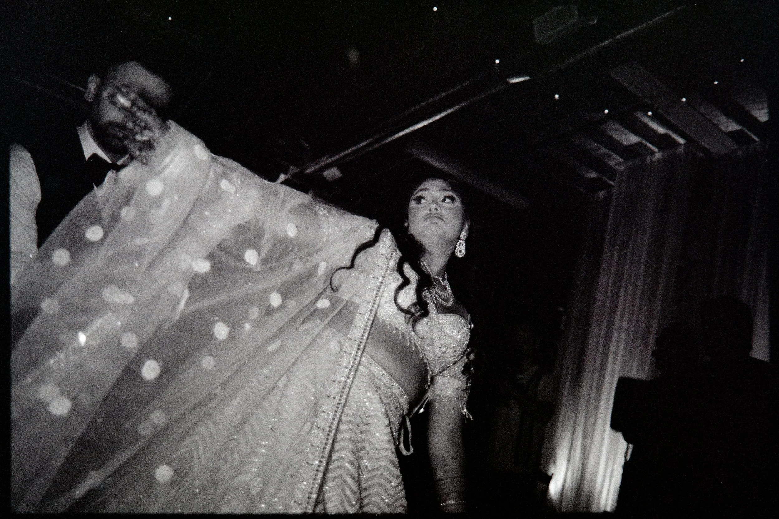 Black and white film photo of an Indian bride in traditional silver and glittery lehenga attire dancing at reception, with the groom partially visible beside her, in a dimly lit setting with curtains.