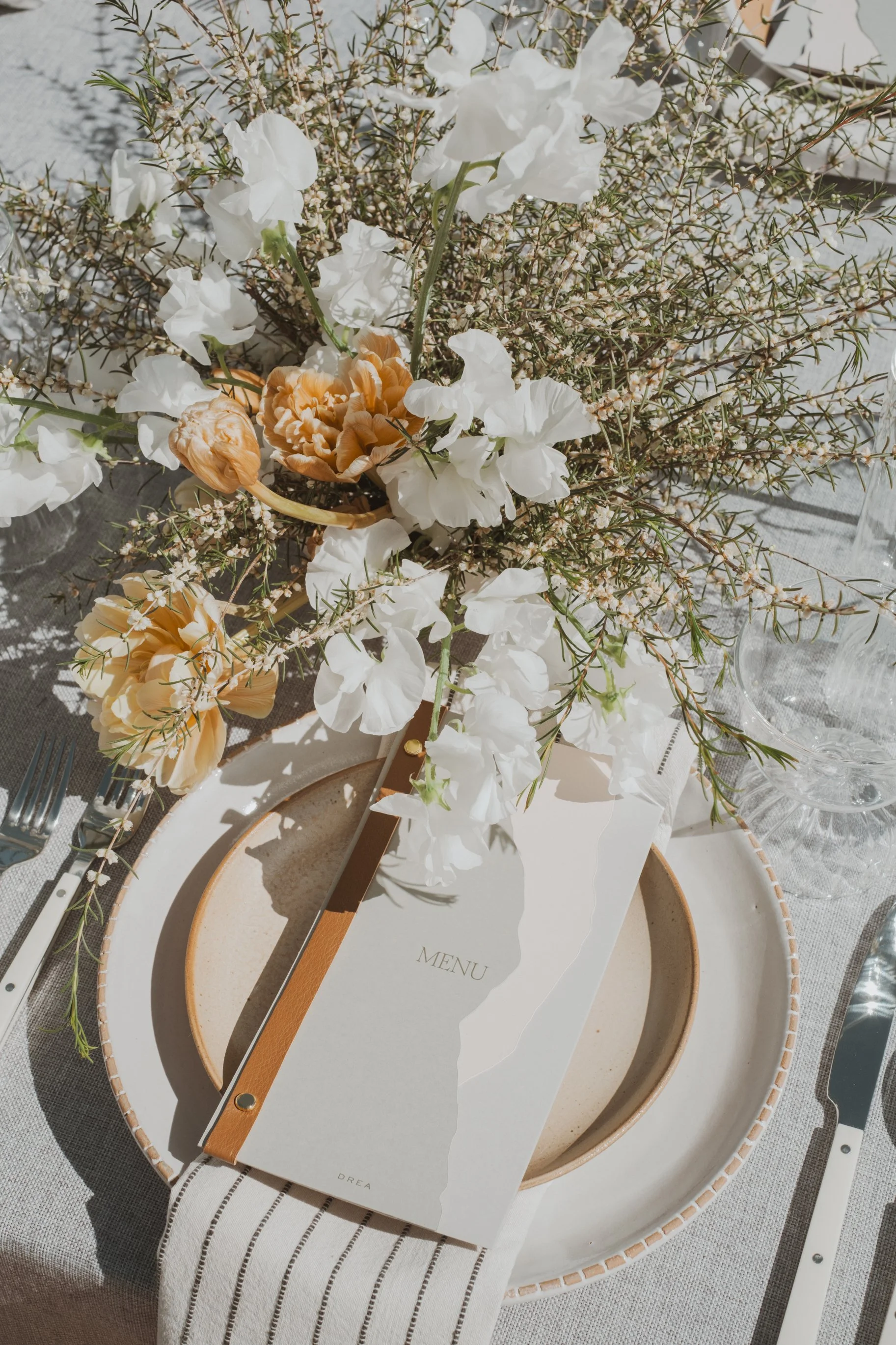 A table setting with a beige plate, a menu card, floral centerpiece, and cutlery arranged on a linen tablecloth.