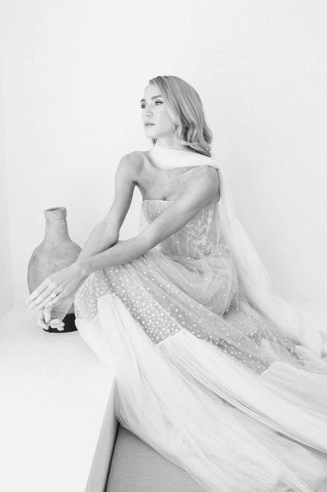 Black and white photo of a woman in a Liz Martinez, embellished dress sitting on a white surface with a clay vase beside her, looking to the side.