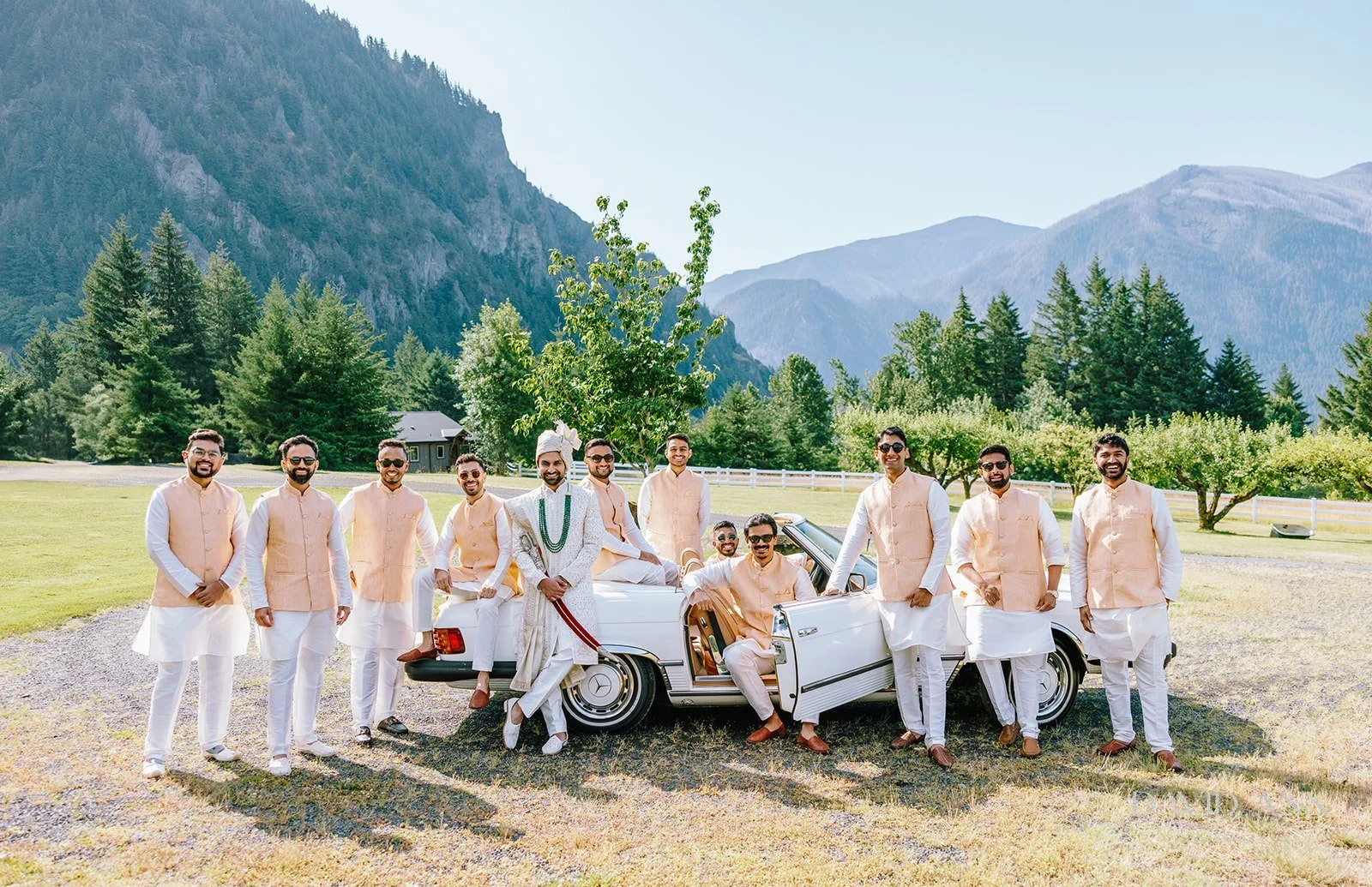 Groom and groomsmen in traditional Indian attire celebrating outdoors with a vintage white car, mountains, and trees in the background.