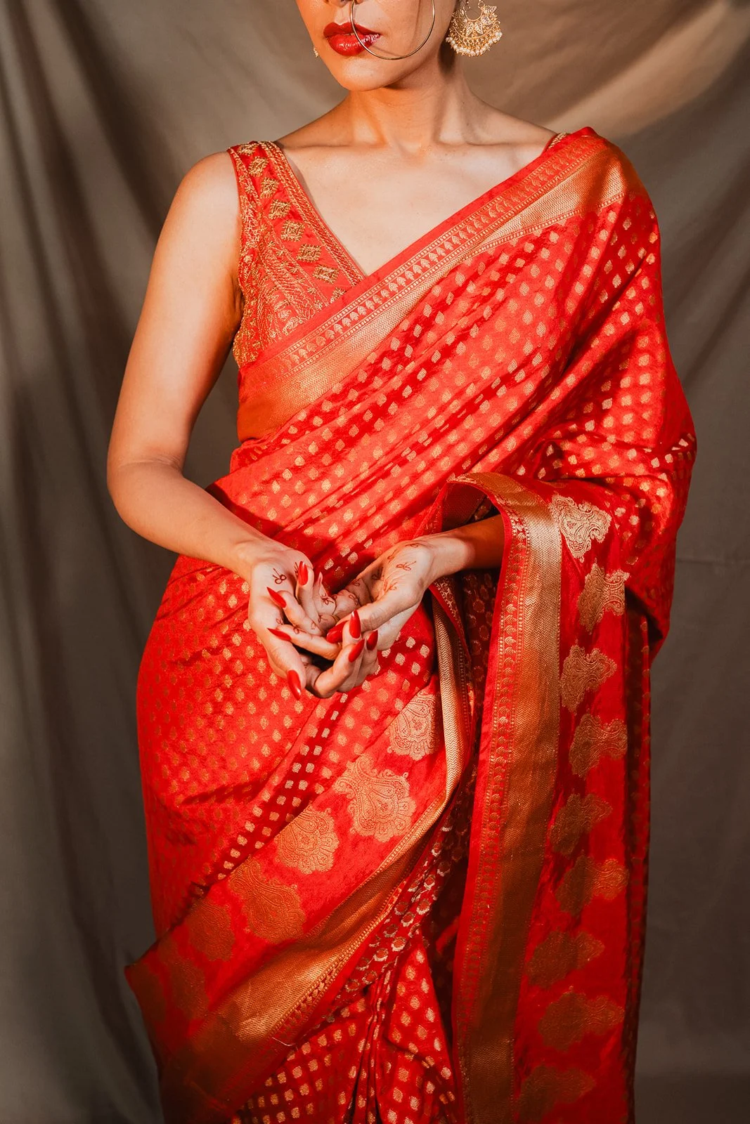 A woman wearing a red sari with gold accents and embroidery, holding her hands in front of her with henna designs on her fingers.