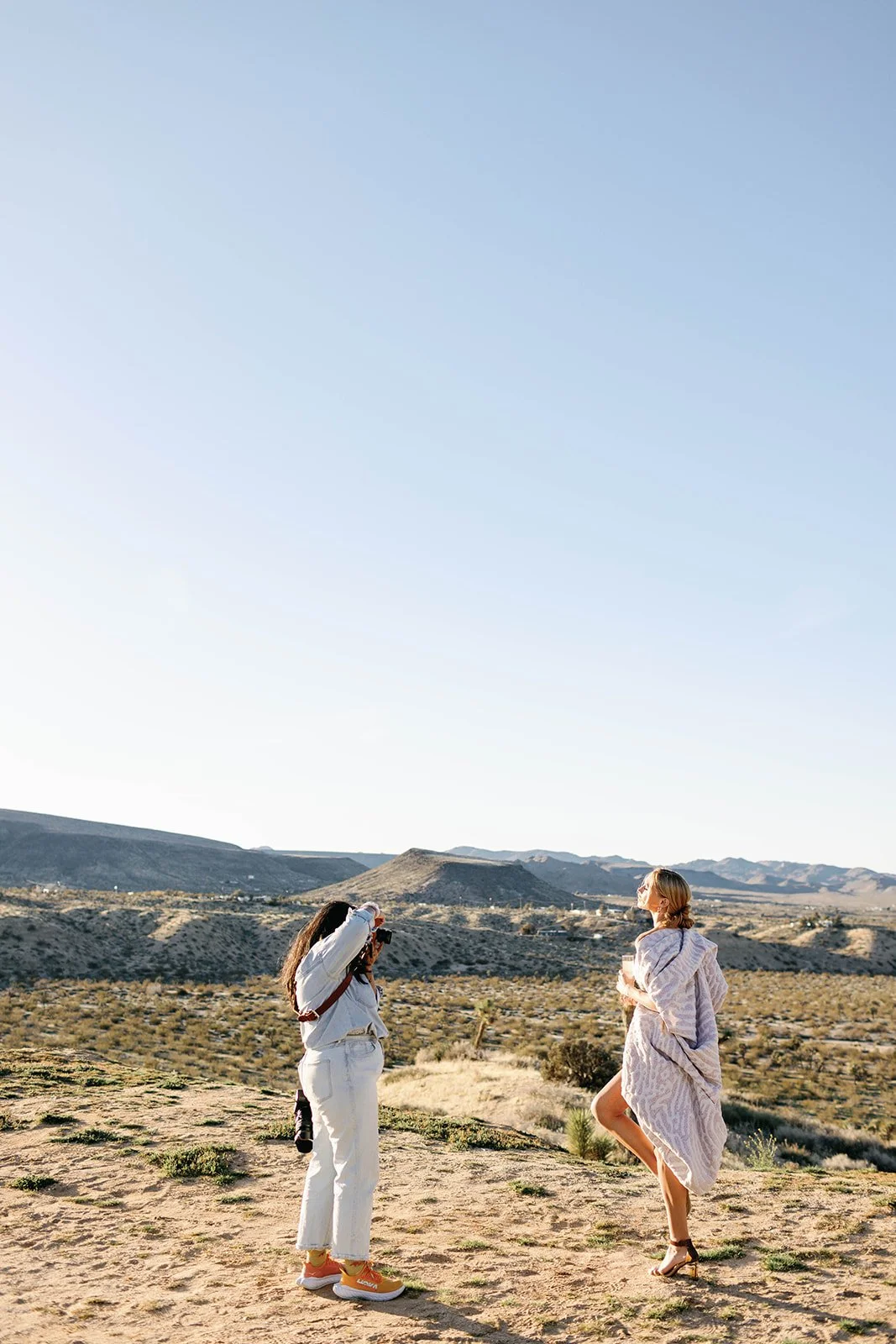 Two women in a desert landscape, one taking a photo of the other who is posing in a dress with a mountainous background.