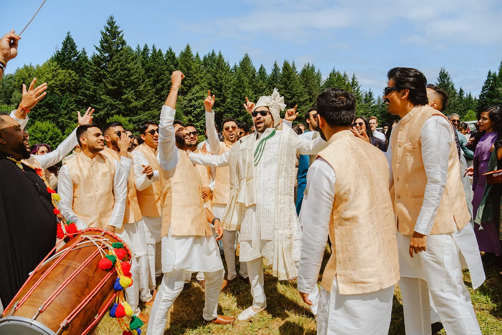 Group of happy people in traditional Indian wedding attire celebrating outdoors with music and dancing. The groom, wearing sunglasses and a cream outfit, is in the center with arms raised. The guests are clapping and smiling, with a decorated drum visible on the left. Trees and a blue sky form the background.