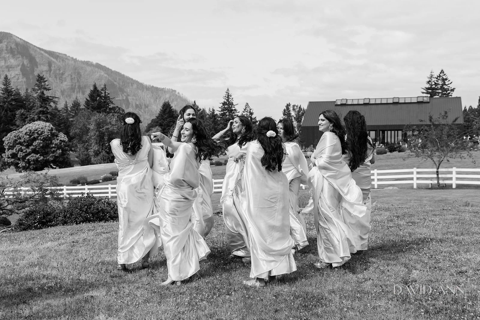 A group of women in wedding dresses dancing outdoors on a lawn with mountains and trees in the background.