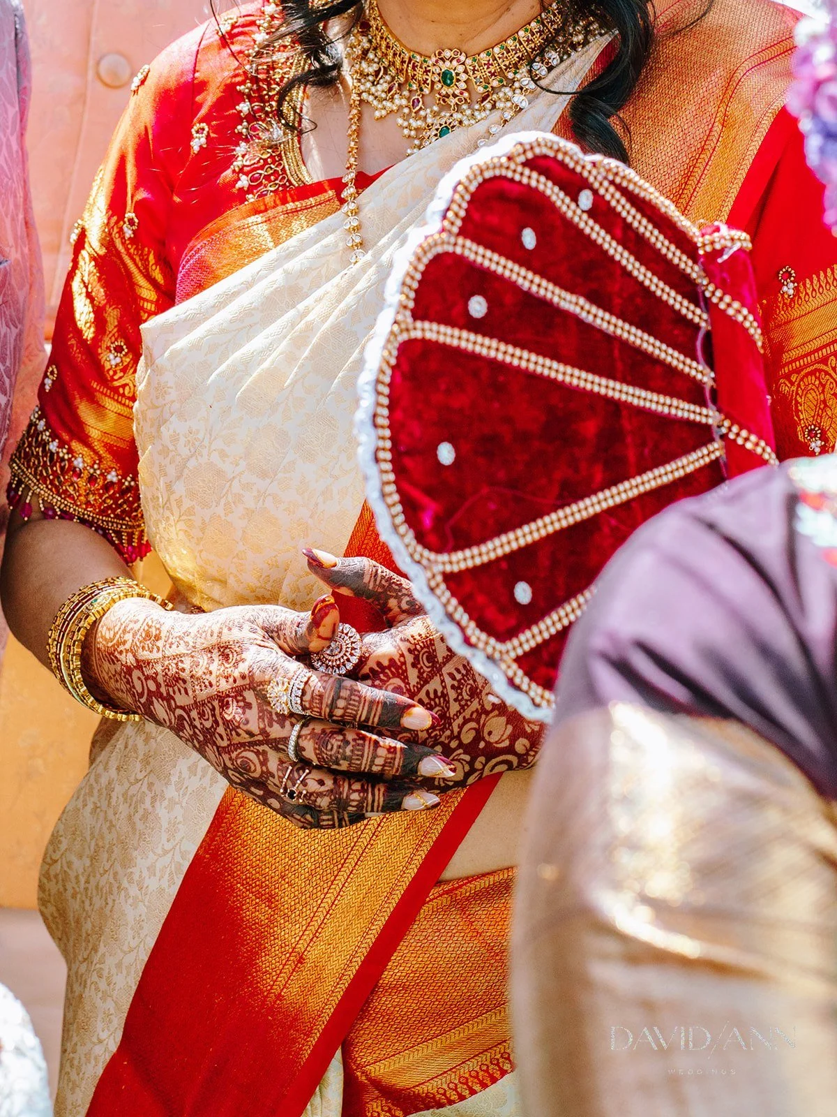 A woman dressed in traditional Indian attire, wearing an intricate necklace, bangles, and rings, holding a red and gold embellished fan or decorative accessory. Her hands are decorated with henna.