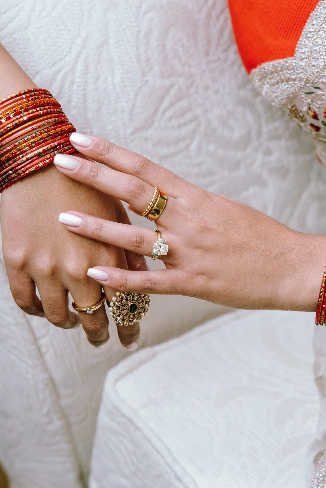 Close-up of hands adorned with colorful bangles and various gold rings, including a large statement ring with a green stone and multiple small gemstones, against a background of traditional Indian clothing.