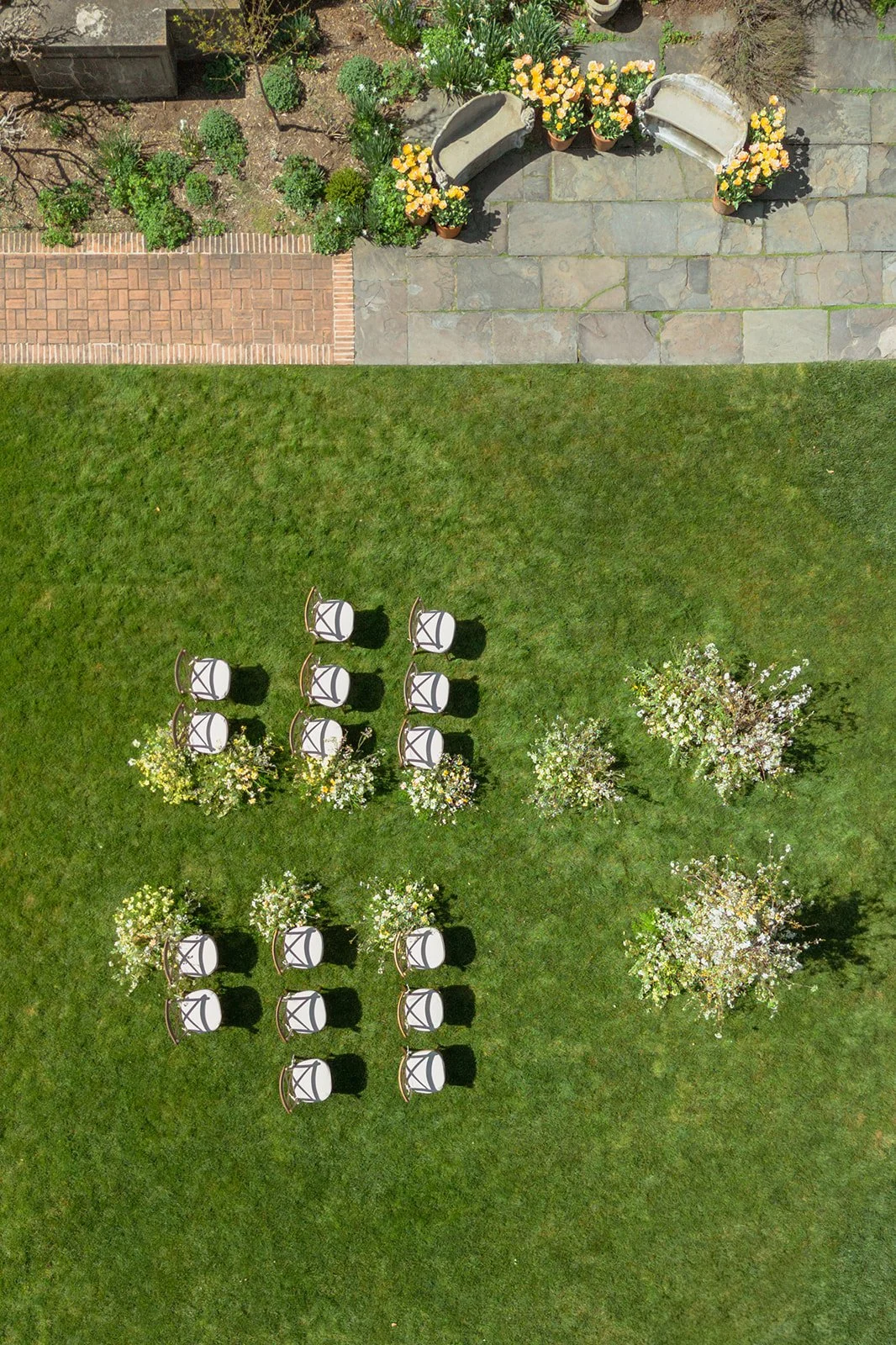 Top-down view of a grassy lawn with arranged chairs and flowering bushes, adjacent to a stone and brick patio area with potted plants and garden benches.