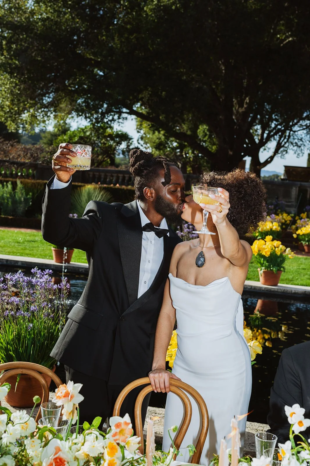 A couple dressed in bridal attire sharing a kiss while holding glasses of champagne at their reception table held in Filoli Gardens, in California.