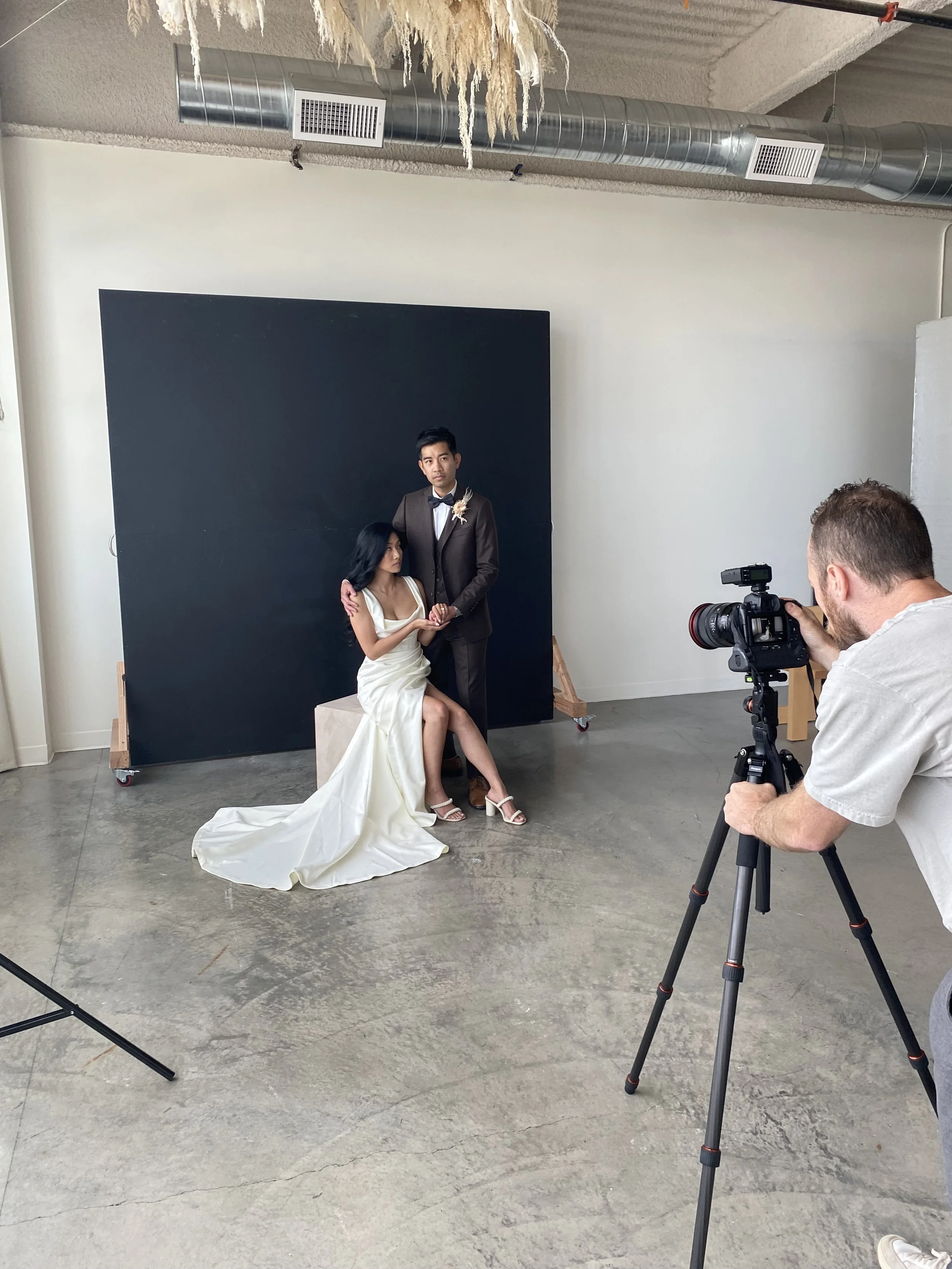 A couple is posing for a wedding photo shoot in a studio. The woman is sitting on a block, wearing a white wedding dress and high heels, while the man stands beside her in a dark suit with a bowtie. A photographer is taking their picture with a professional camera, and a black backdrop is behind them.