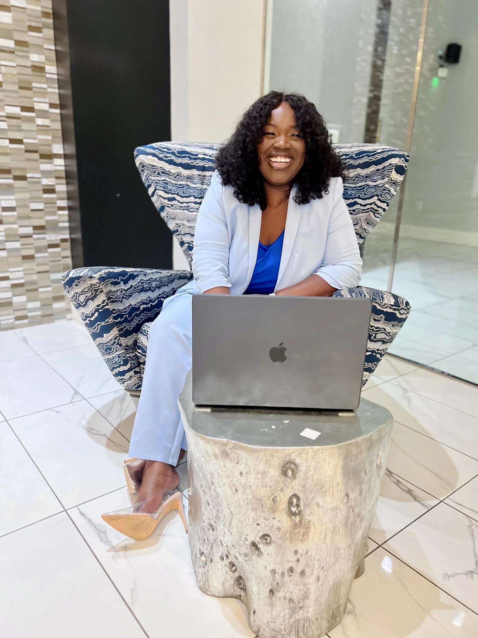 A woman with curly black hair and a bright smile sitting in a modern lobby area with a laptop in front of her.