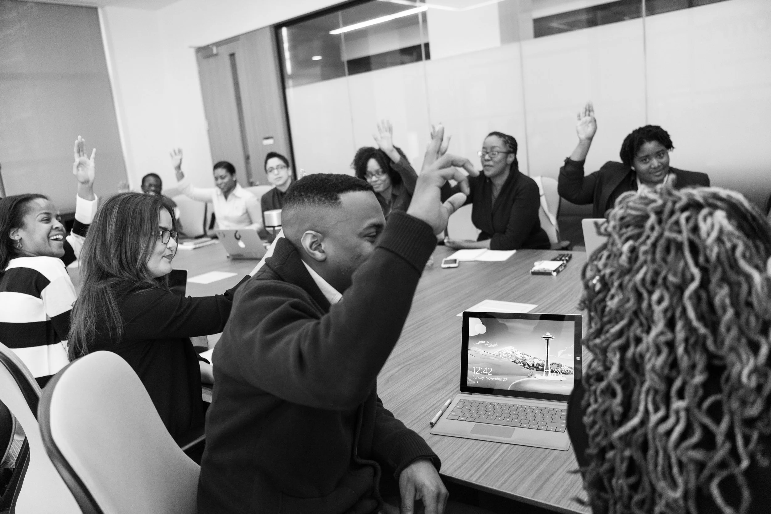 A diverse group of people in a business meeting, some raising hands, sitting around a large conference table with laptops, notebooks, and a tablet showing the Seattle Space Needle, in a modern office.